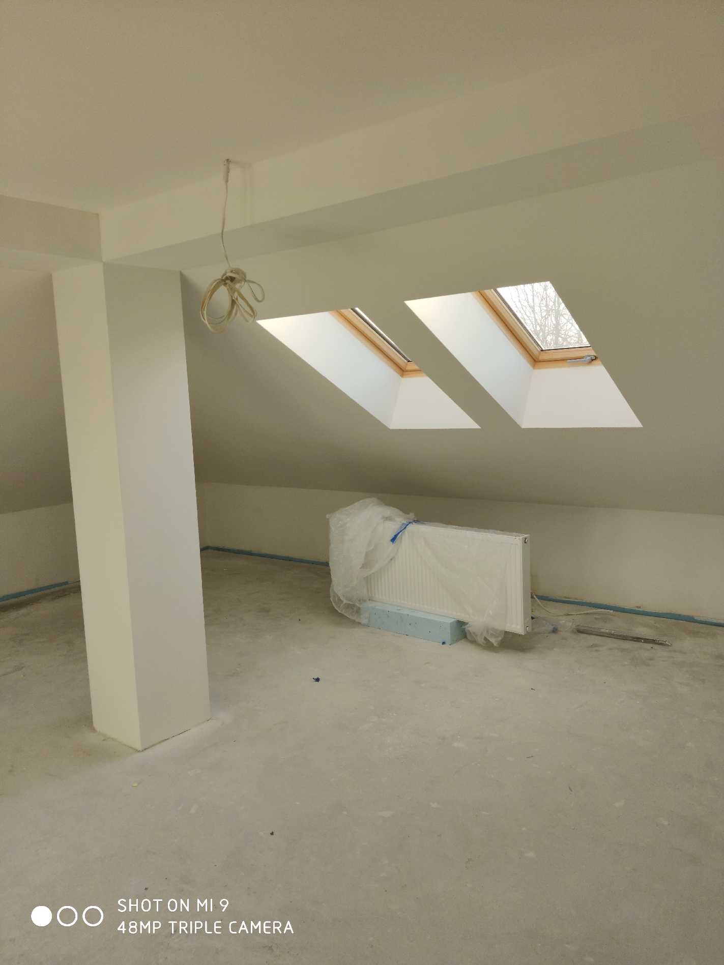 Poddasze po świeżym malowaniu na biało, z dwoma oknami dachowymi i grzejnikiem zabezpieczonym folią, widoczny surowy beton na podłodze i wiszące kable elektryczne.