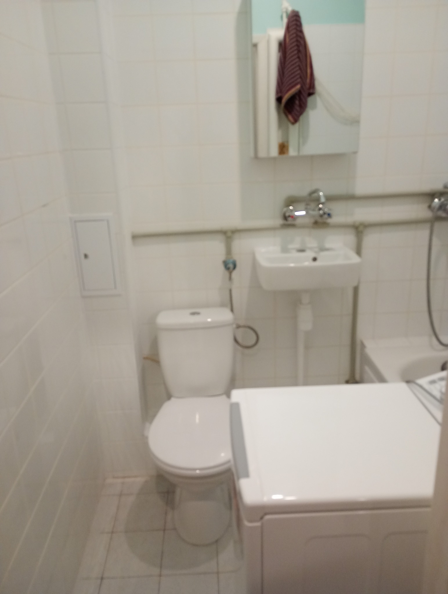 Standardowa, biała łazienka z toaletą, umywalką, lustrem z wiszącym ręcznikiem i pralką, wykończona białymi płytkami.