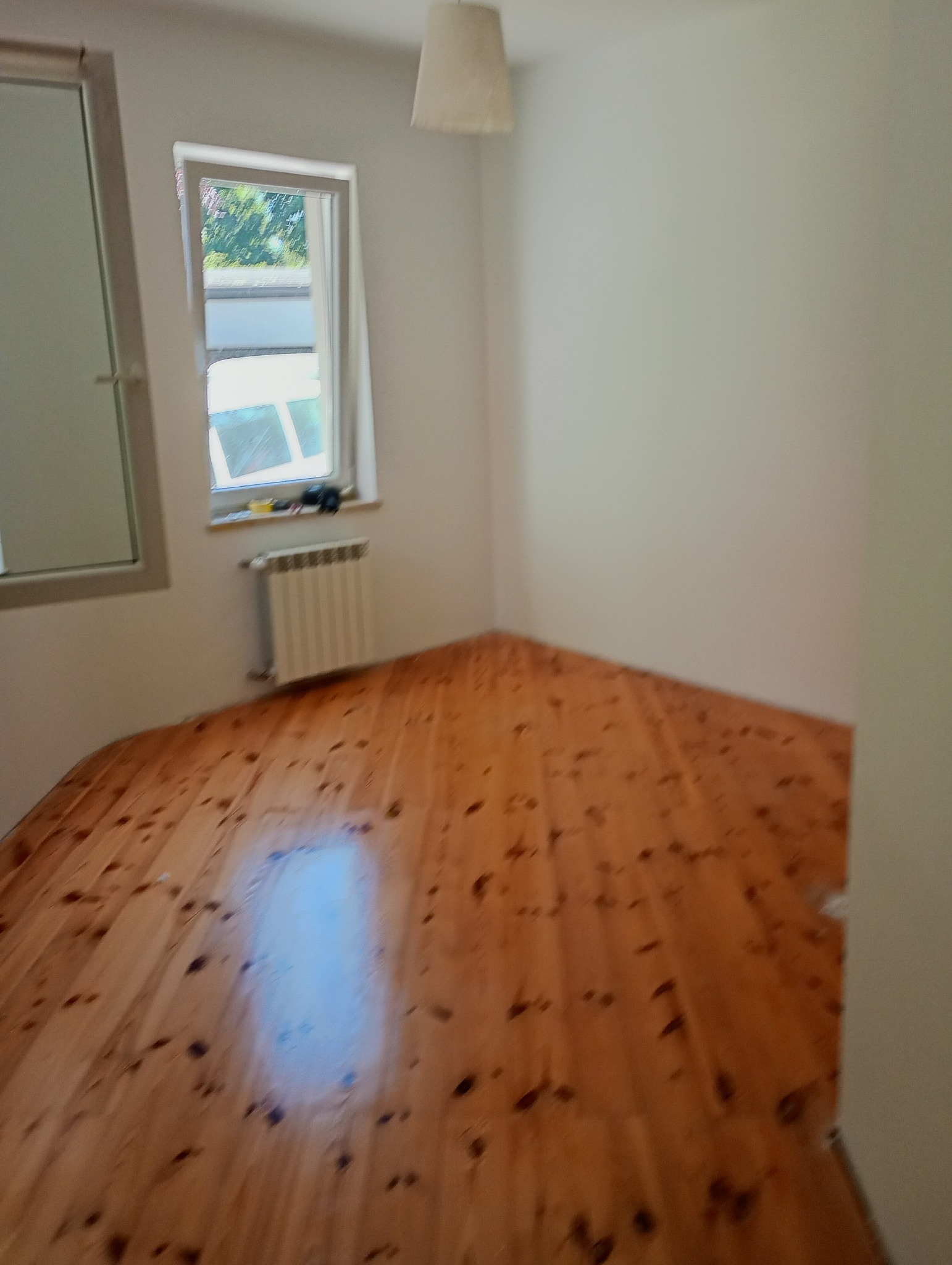 Pomalowane na biało wnętrze pokoju z drewnianą podłogą i grzejnikiem pod oknem. Widoczna lampa wisząca. Świeżo odmalowane ściany.