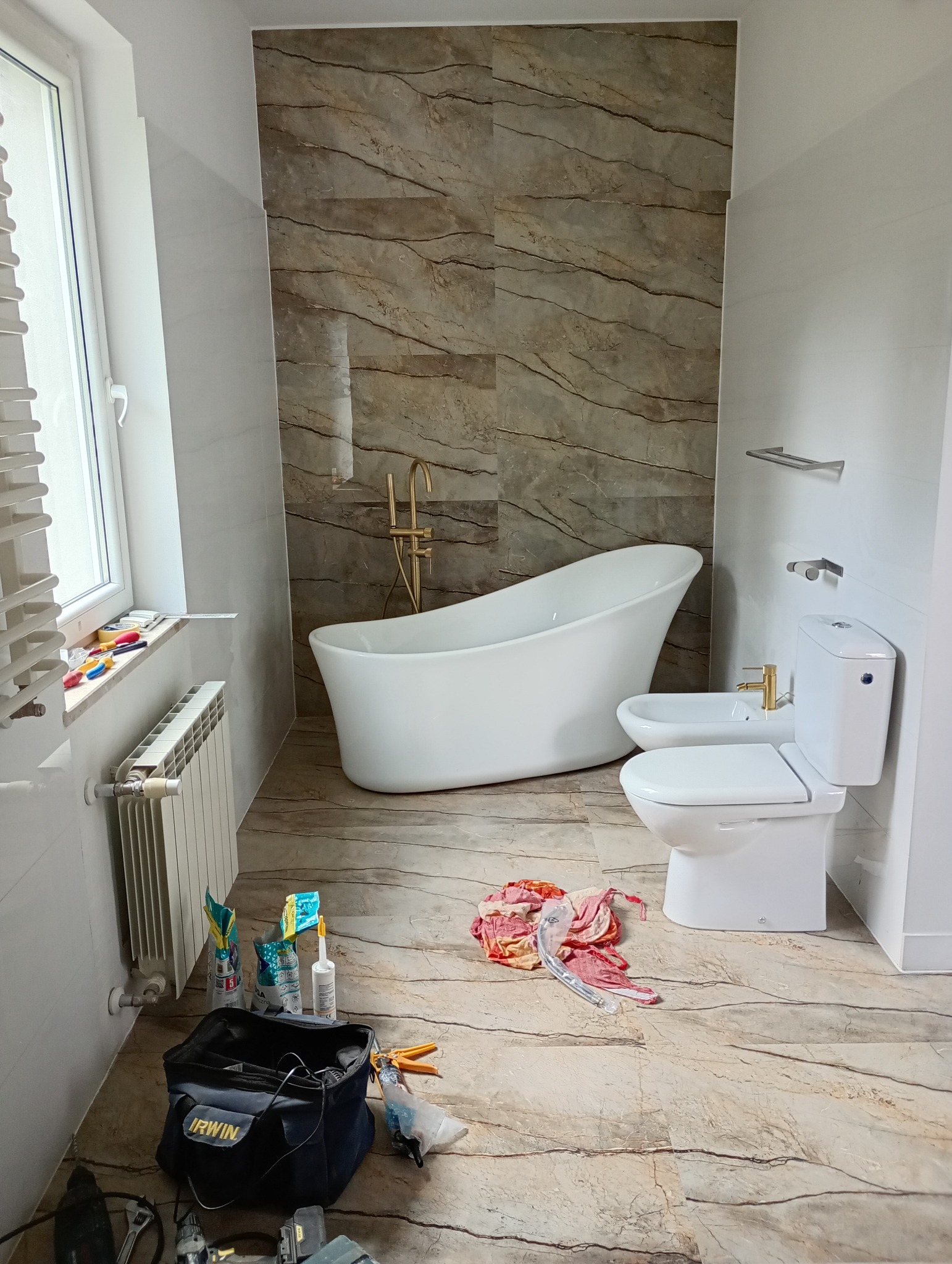 Wykończona łazienka z wolnostojącą wanną, bidetem i toaletą. Na podłodze i ścianie imitacja marmuru. Widoczne narzędzia i akcesoria instalacyjne.