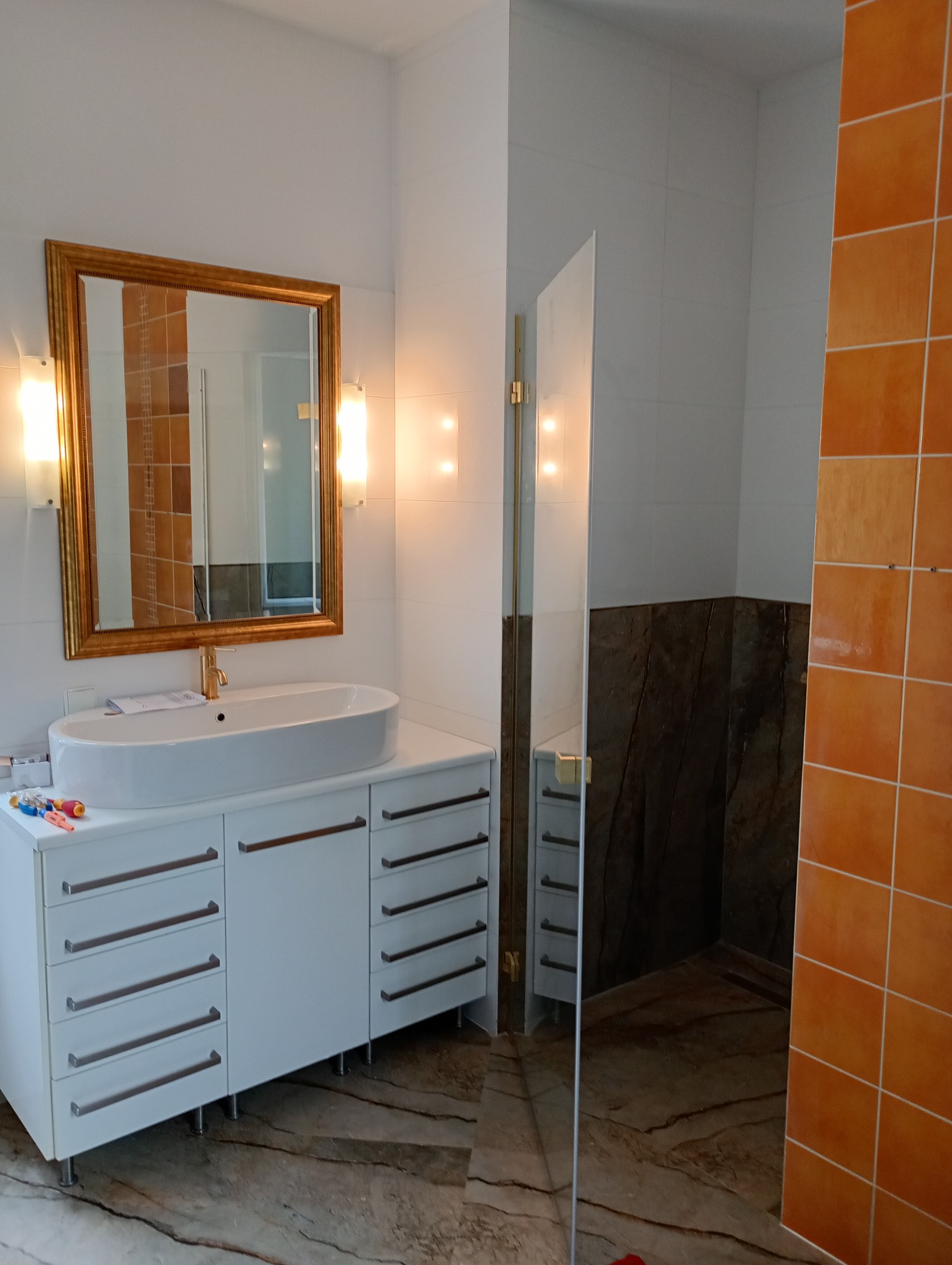 Wykończona łazienka z marmurową podłogą, białą szafką pod umywalką i złotą armaturą. W tle kabina prysznicowa z brązowymi płytkami i ścianą z pomarańczowych płytek.