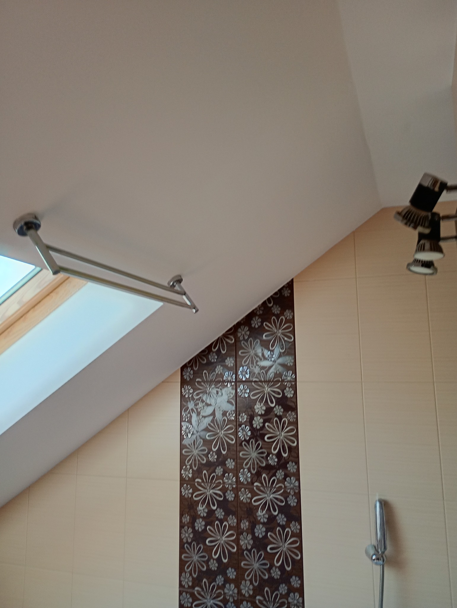 Ukończona łazienka na poddaszu: beżowe płytki, dekoracyjny pas z motywem kwiatowym, okno dachowe i oświetlenie punktowe. Widok z dołu.