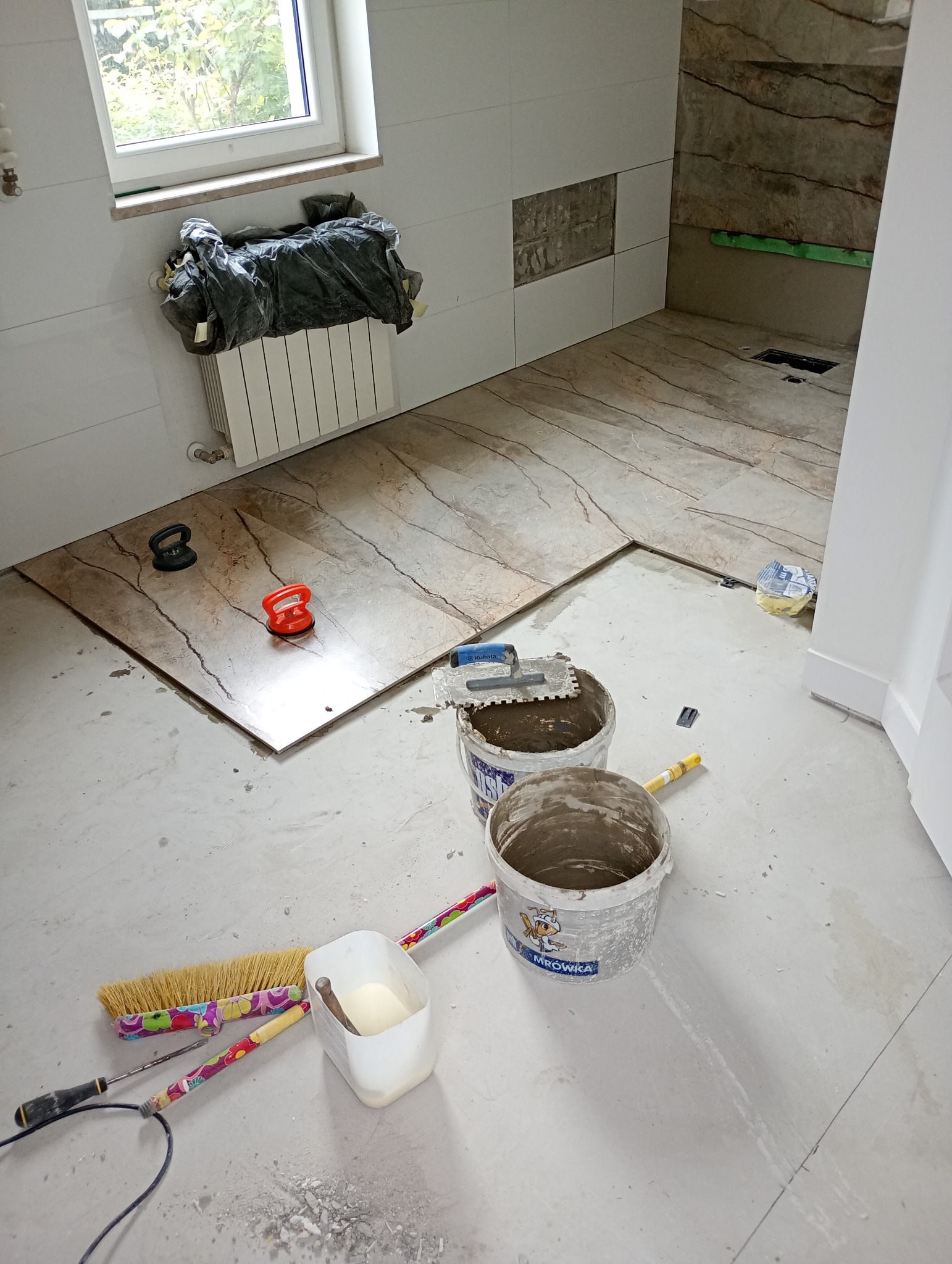 Układanie płytek podłogowych w łazience w trakcie remontu. Widoczne narzędzia, wiaderka z zaprawą i częściowo ułożone płytki imitujące marmur.