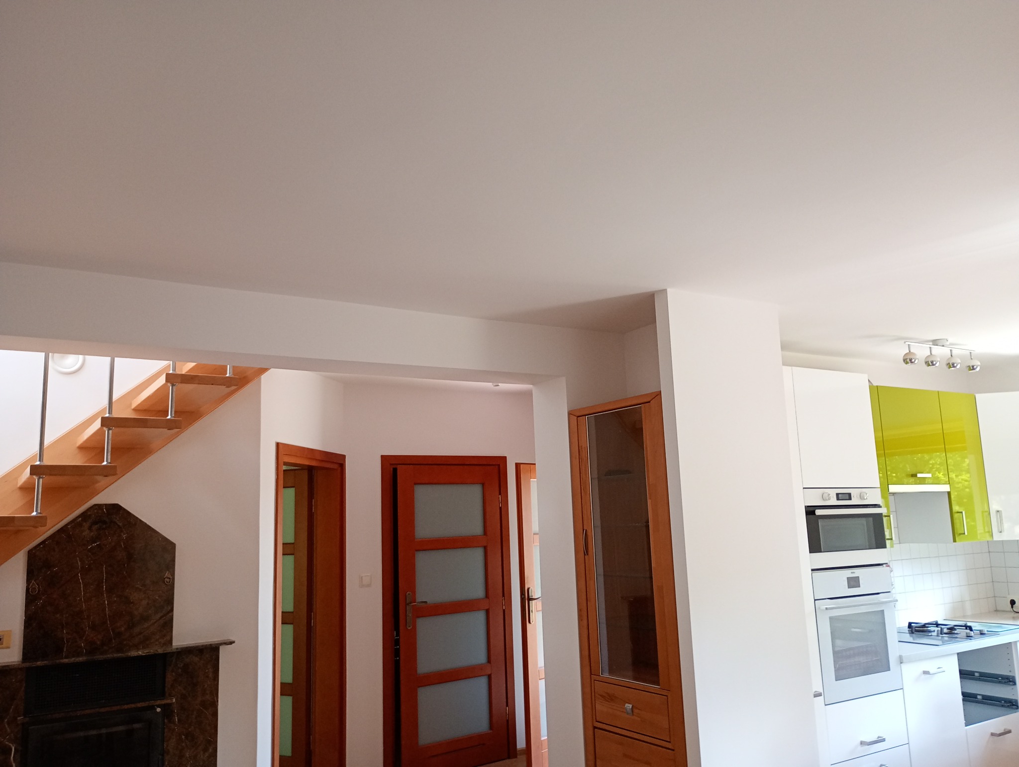 Jasne wnętrze domu po remoncie: drewniane schody, dębowe drzwi z przeszkleniami i nowoczesna kuchnia z zielonymi akcentami. Widoczny fragment kominka z marmuru.