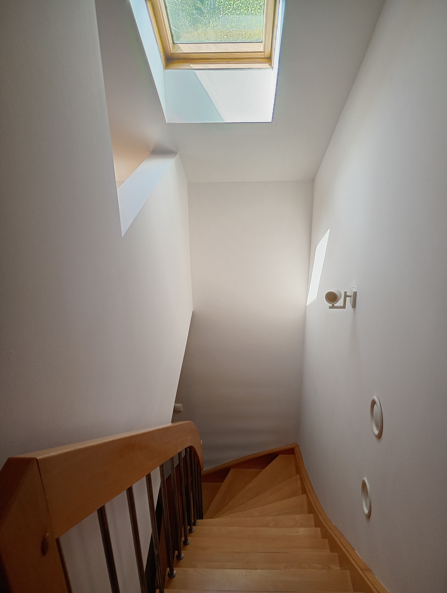 Jasne schody z drewnianymi poręczami w nowoczesnym domu. Widoczne okno dachowe wpuszczające światło. Ściany pomalowane na biało. Minimalistyczne oświetlenie.