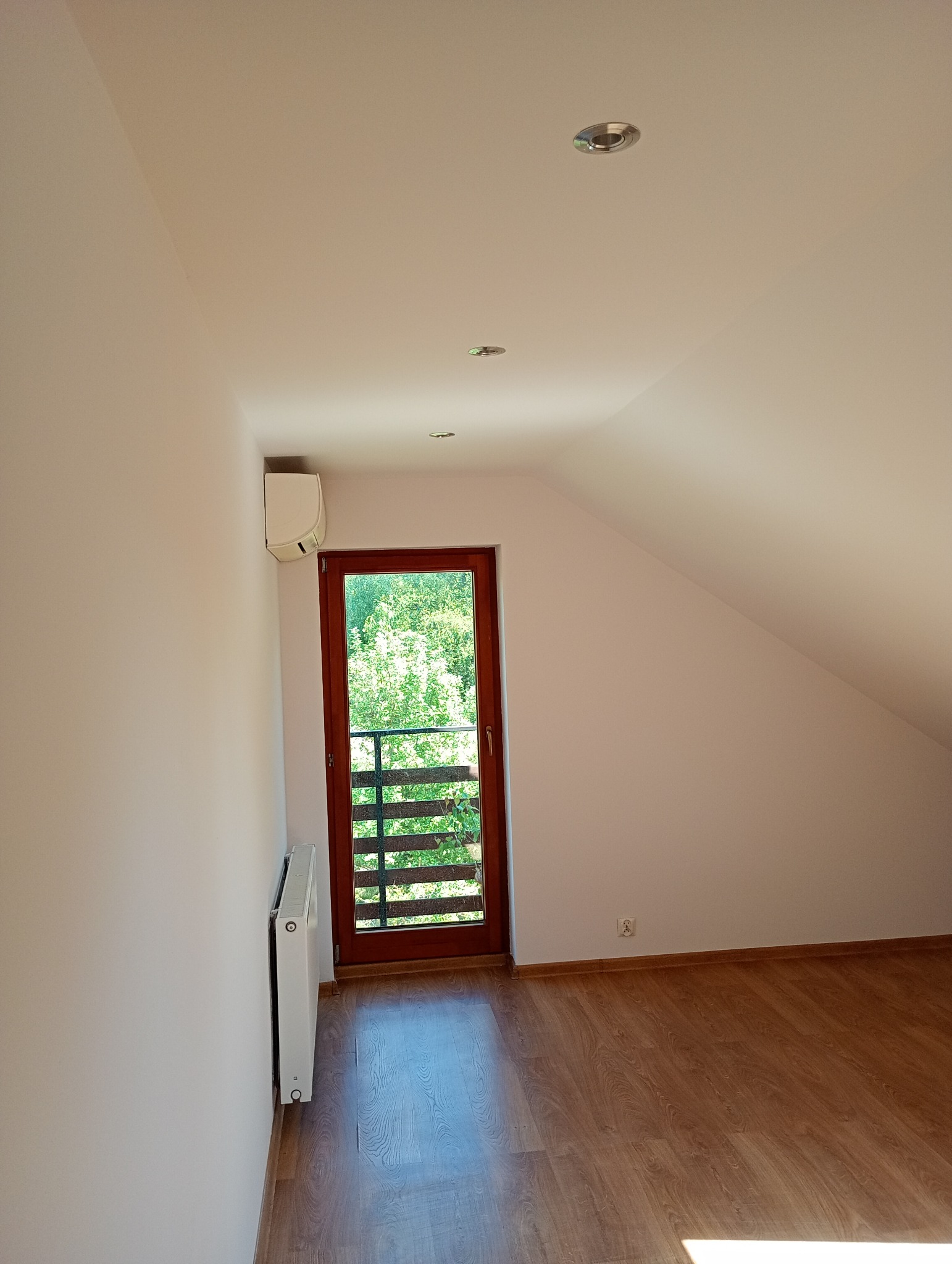 Poddasze po remoncie: świeżo pomalowane białe ściany, drewniana podłoga, drzwi balkonowe z widokiem na zieleń. Zamontowany grzejnik i klimatyzator.