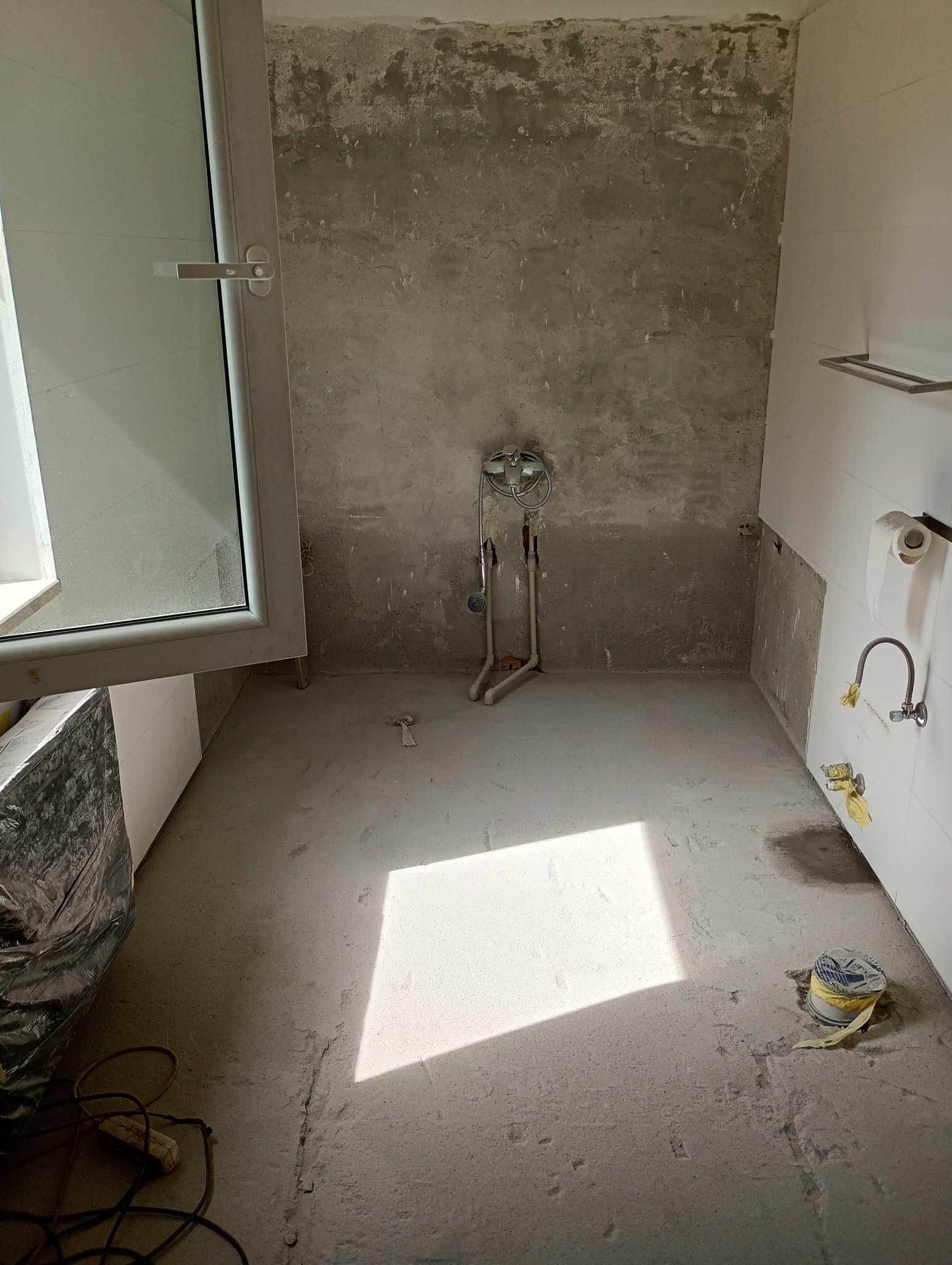 Wnętrze łazienki w trakcie remontu: surowe ściany, instalacja wodna, otwarte okno, narzędzia i materiały budowlane na podłodze. Widok na etap prac przygotowawczych.