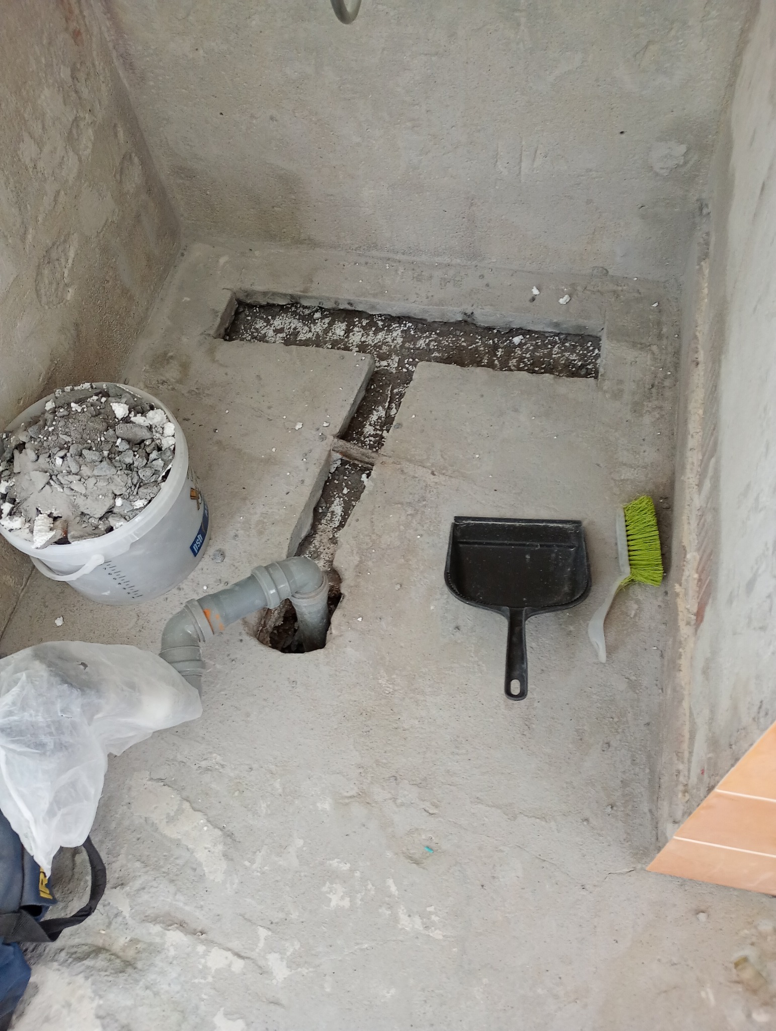 Fragment podłogi w trakcie remontu: widoczna rura kanalizacyjna, wiadro z gruzem, szpachelka i szczotka. Podłoże betonowe z wyciętymi fragmentami.