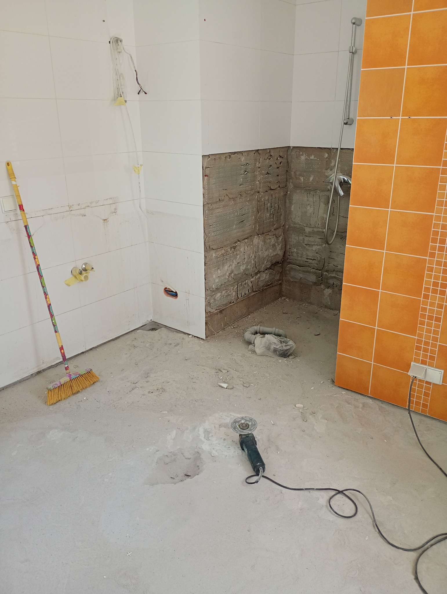 Remont łazienki w trakcie: częściowo zdjęte płytki, widoczne instalacje, narzędzia (szlifierka kątowa) i miotła. Pomarańczowe płytki na ścianie.