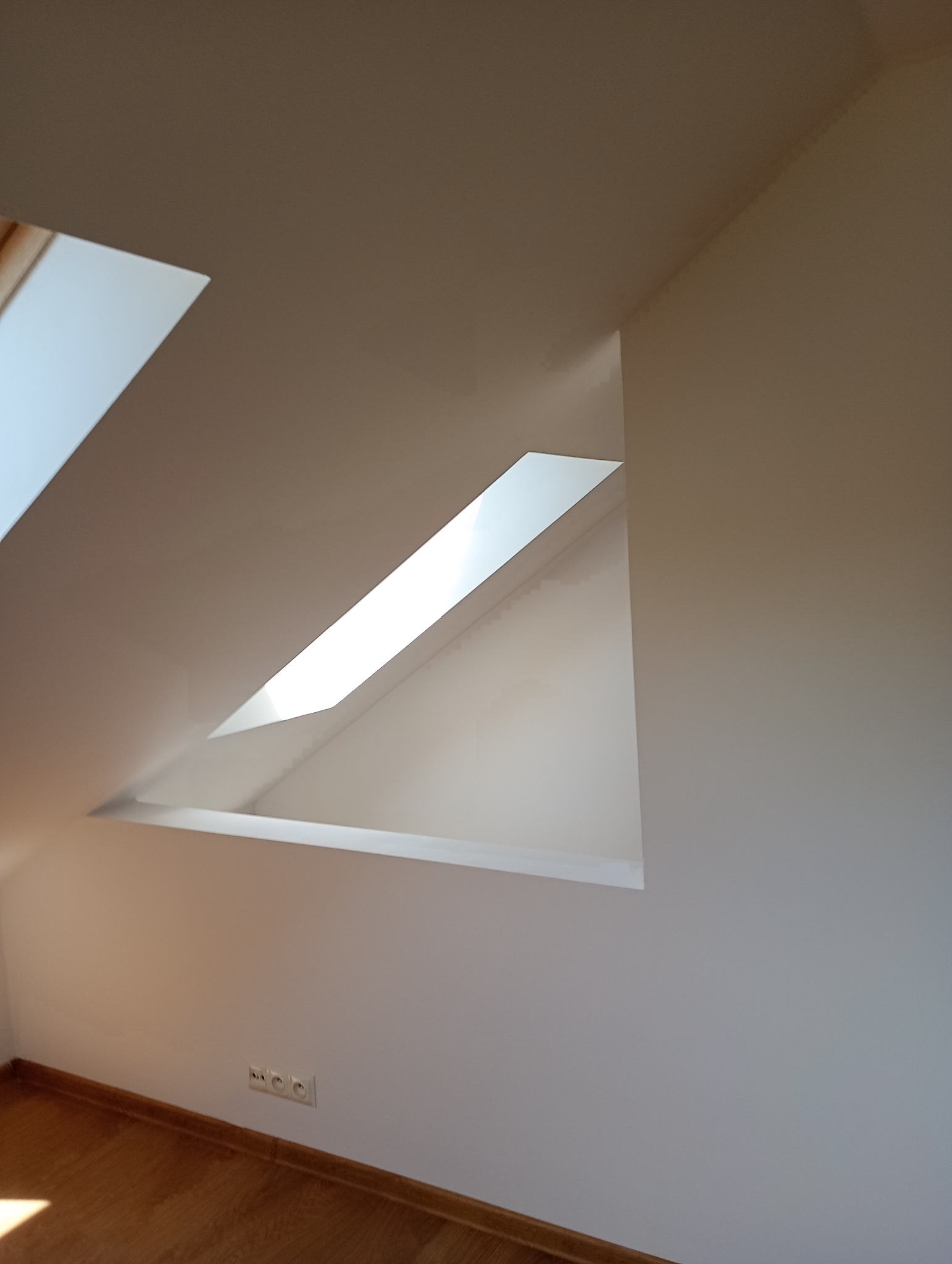 Białe ściany i drewniana podłoga poddasza z oknem dachowym w kształcie trapezu, wnętrze po malowaniu. Widoczne gniazdka elektryczne.