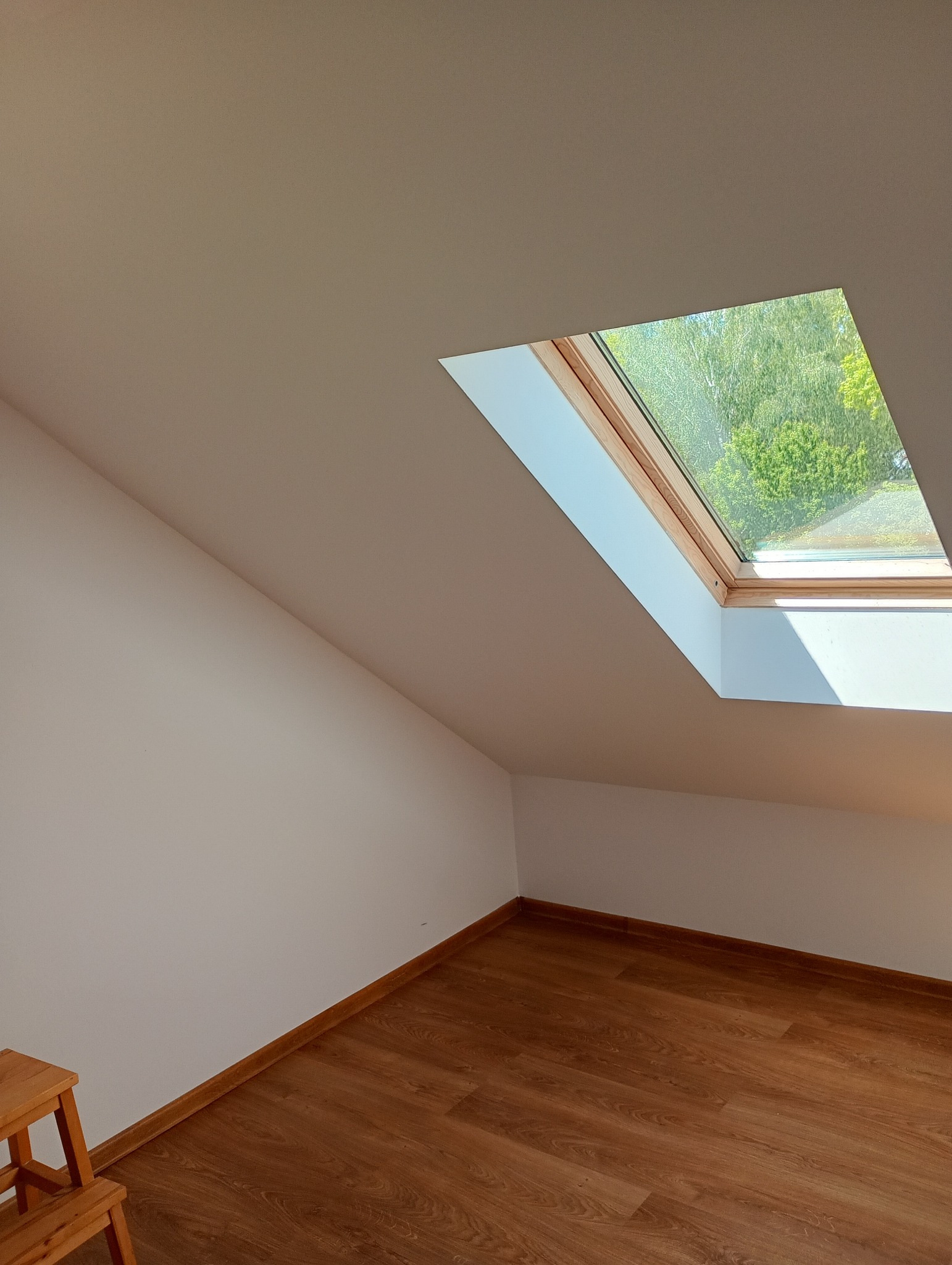 Poddasze z oknem dachowym i drewnianą podłogą. Widoczny fragment drewnianego stołka malarskiego. Ściany pomalowane na jasny kolor.