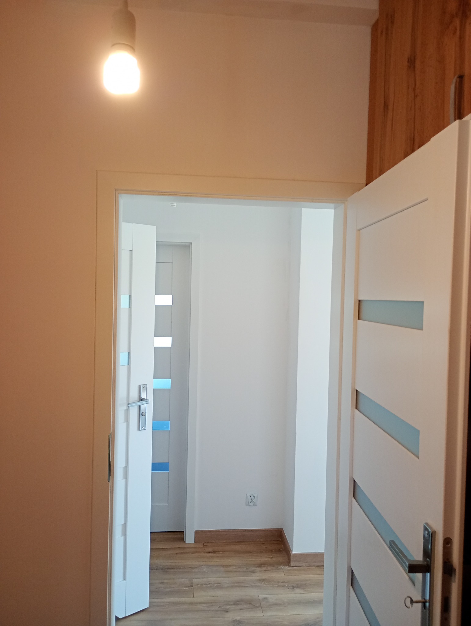 Białe, częściowo otwarte drzwi z przeszkleniami w odcieniach błękitu, widoczne z perspektywy innego pomieszczenia, z drewnianą podłogą i fragmentem drewnianej ściany.