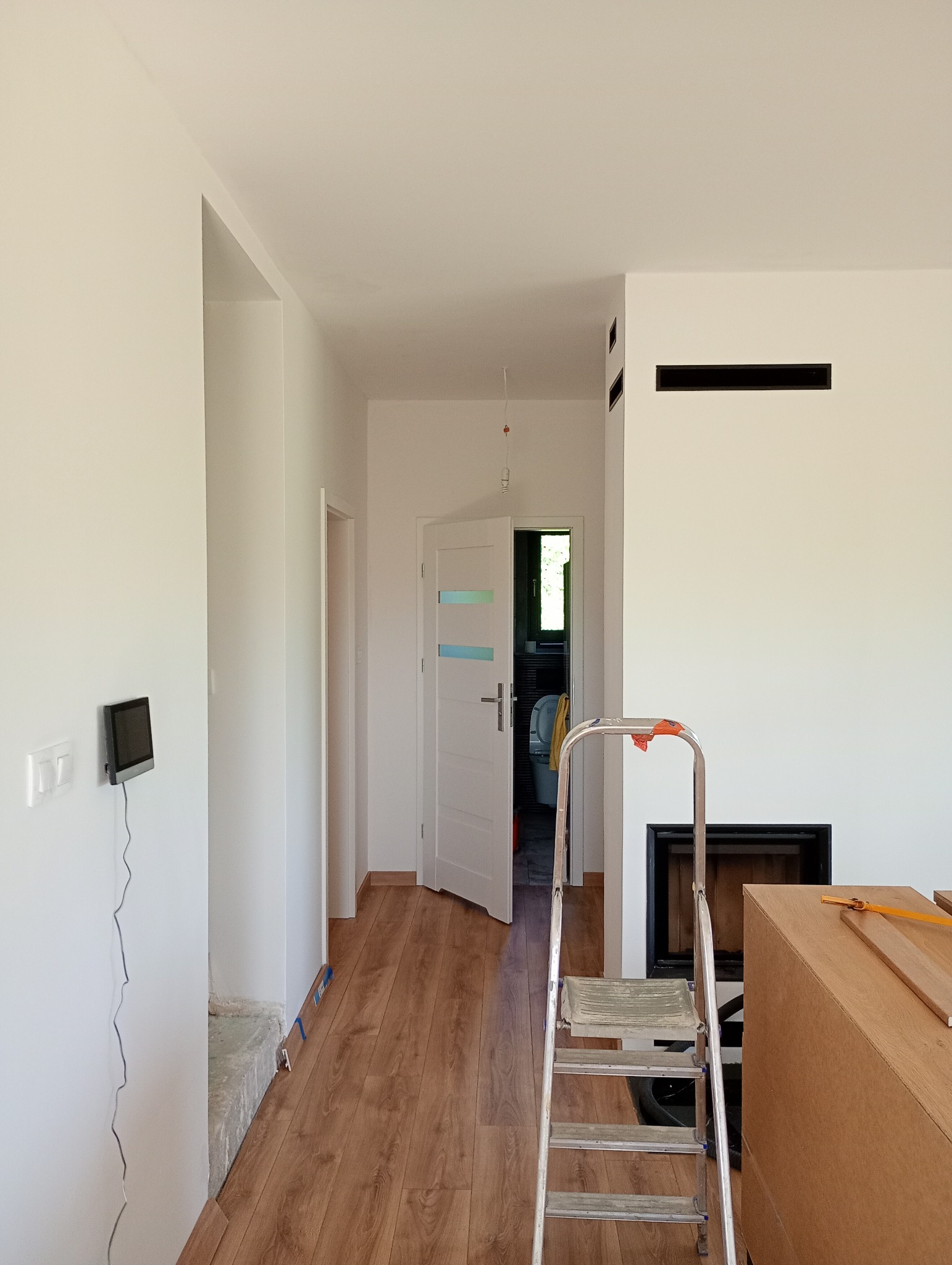 Wnętrze mieszkania w trakcie remontu: białe ściany, panele na podłodze, drabina, widok na otwarte drzwi do łazienki z toaletą. Etap wykończeniowy.