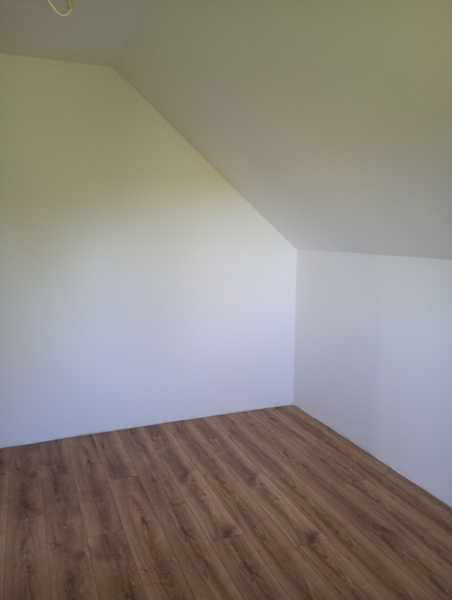 Pomalowane na biało poddasze ze skośnym sufitem i brązowymi panelami podłogowymi. Widok na róg pokoju, sufit i ściany gładkie, jednolite.