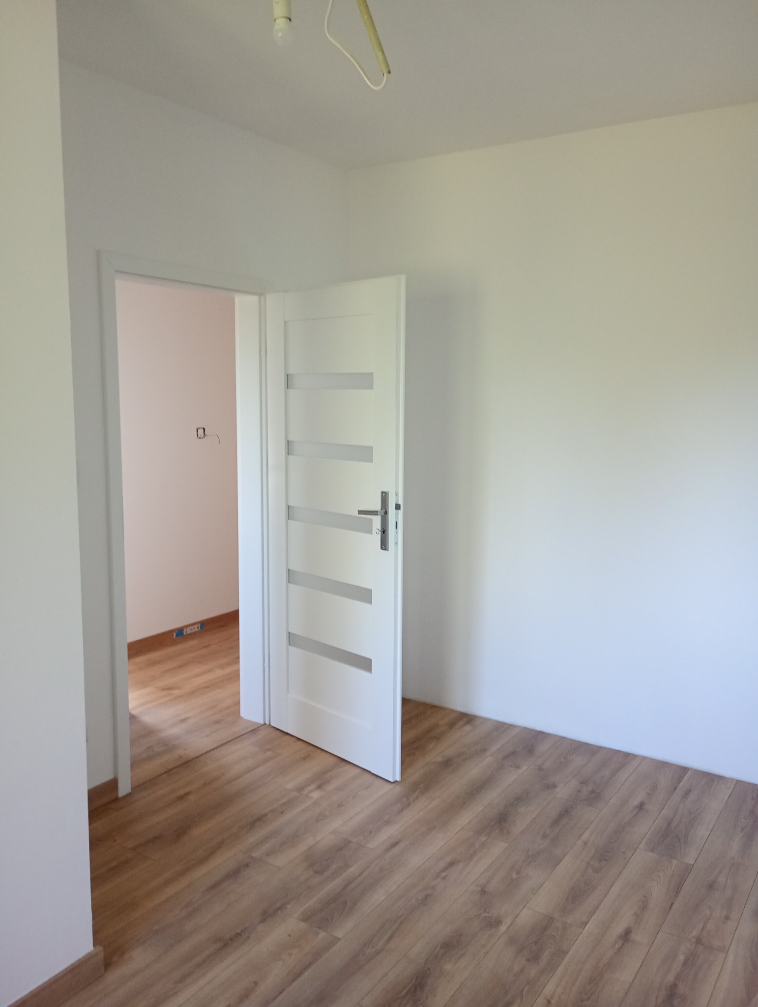 Jasne wnętrze po remoncie: świeżo pomalowane ściany, drewnopodobna podłoga i białe, częściowo otwarte drzwi z przeszkleniami. Widoczne gniazdko elektryczne.