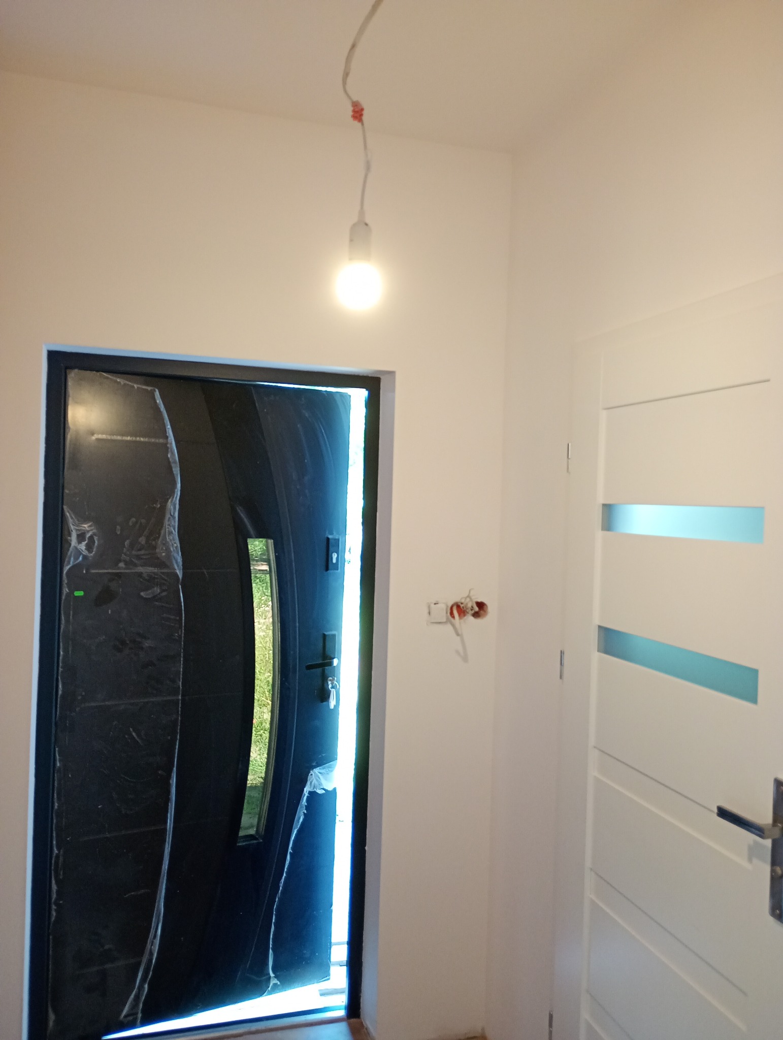 Nowe, czarne drzwi wejściowe, jeszcze zabezpieczone folią ochronną, w jasnym wnętrzu. Widoczna instalacja elektryczna i białe drzwi wewnętrzne.