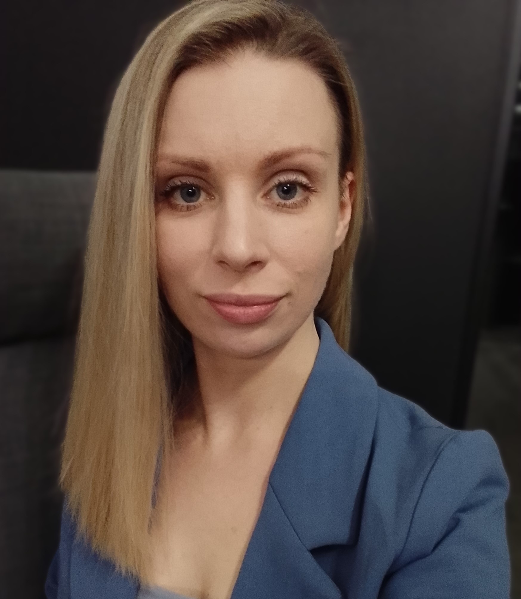 Portret kobiety o jasnych włosach i niebieskich oczach, ubrana w niebieską marynarkę, na ciemnoszarym tle.