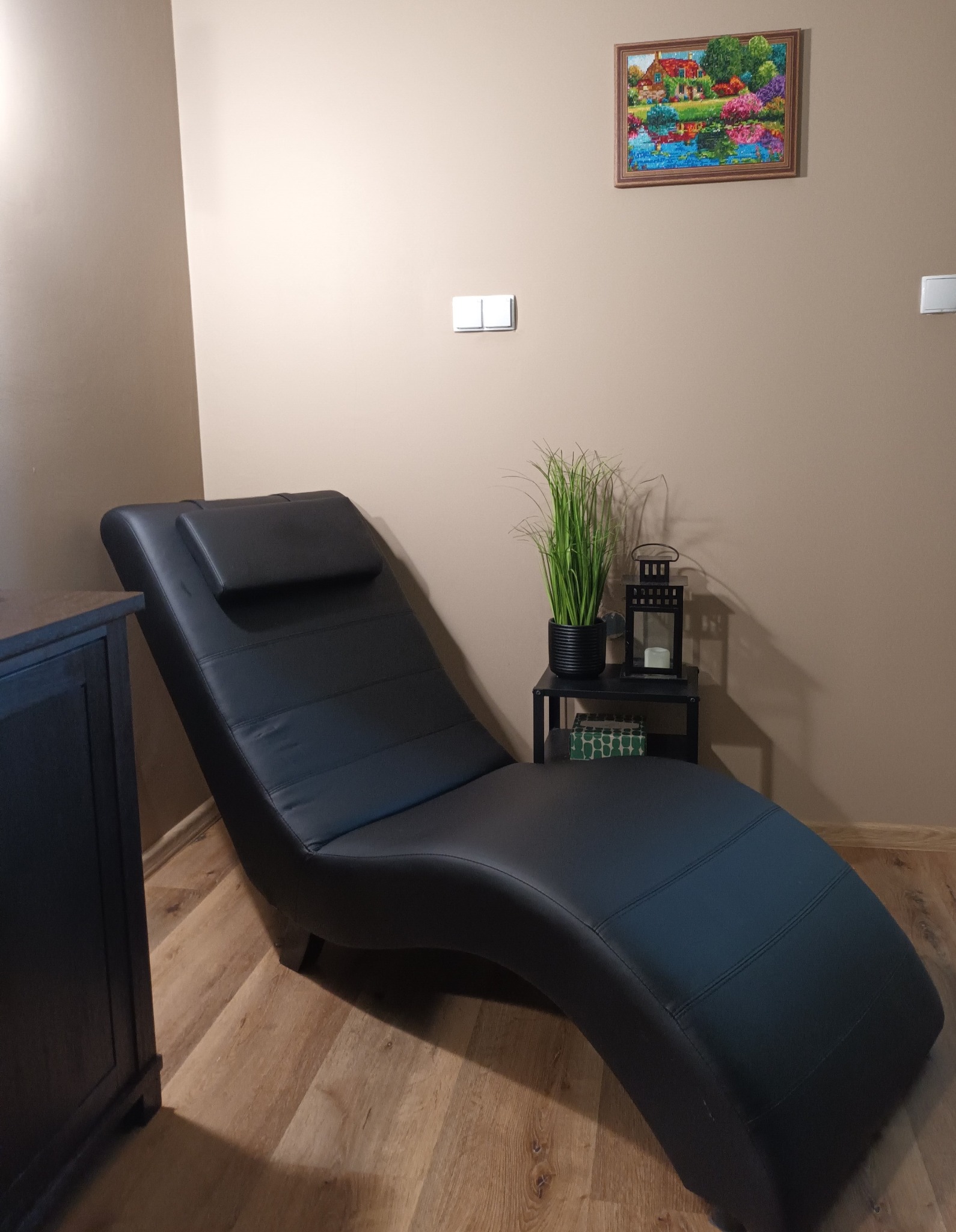 Czarne skórzane leżanka z zagłówkiem w gabinecie, obok czarnej szafki i stolika z lampionem i rośliną w doniczce, na ścianie obrazek w ramce.