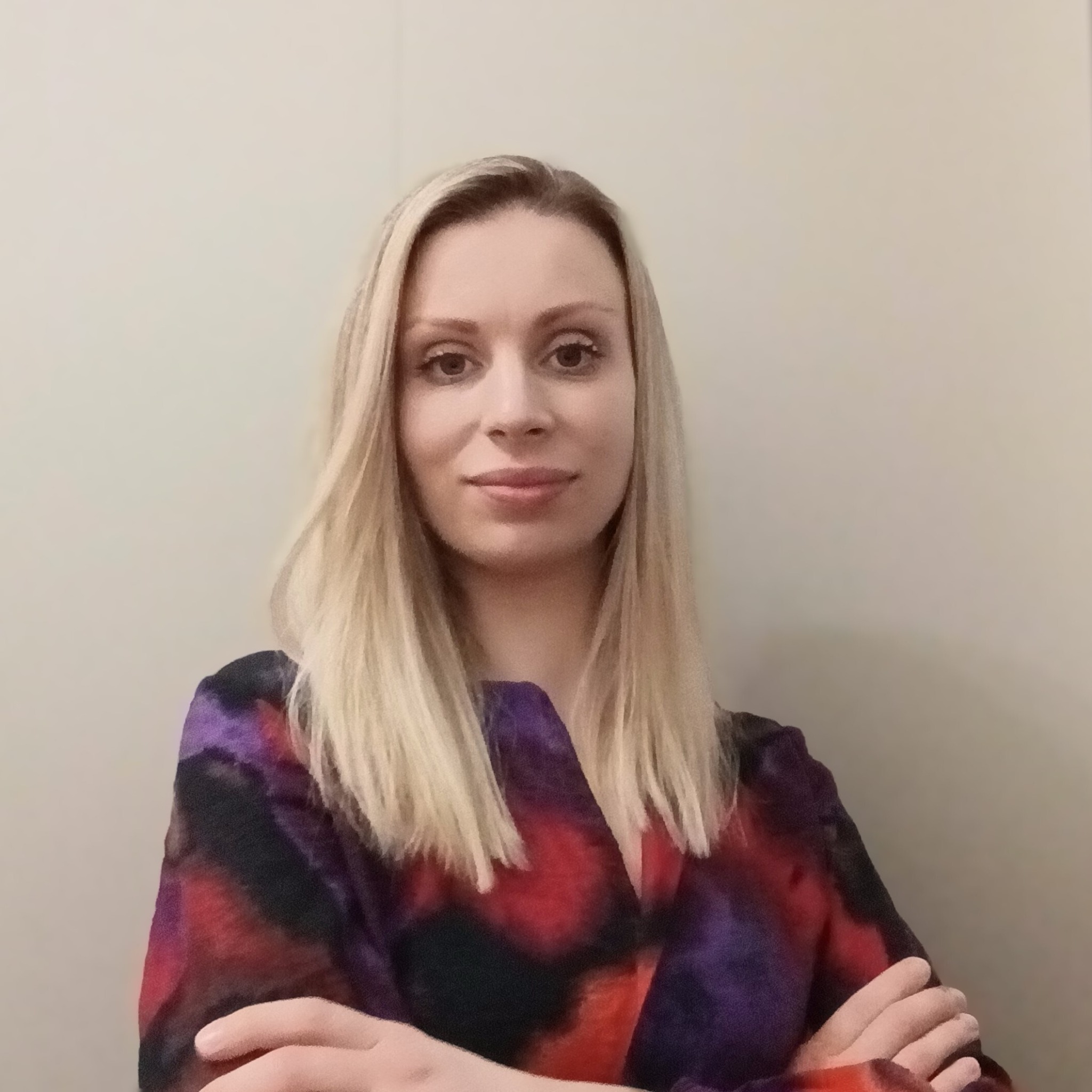 Portret kobiety z blond włosami, ubranej w bluzkę w odcieniach czerwieni, fioletu i czerni, z założonymi rękami na tle jasnej ściany.