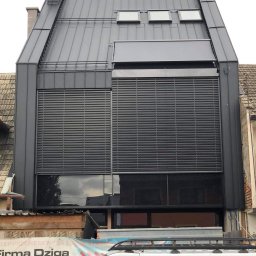 Usługi Ogólnobudowlane - Nowoczesny dom z blachy trapezowej w kolorze grafitowym, okna z żaluzjami zewnętrznymi, dekarz w czerwonej kurtce na dachu, widoczne obróbki blacharskie i komin.