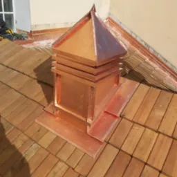 Miedziana, piramidalna obudowa komina na dachu pokrytym drewnianym gontem, widoczny fragment okna i narzędzia dekarskie.