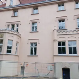 Odnowiona elewacja budynku wielorodzinnego w odcieniach beżu z licznymi oknami o białych ramach, widoczna metalowa siatka zabezpieczająca teren budowy.