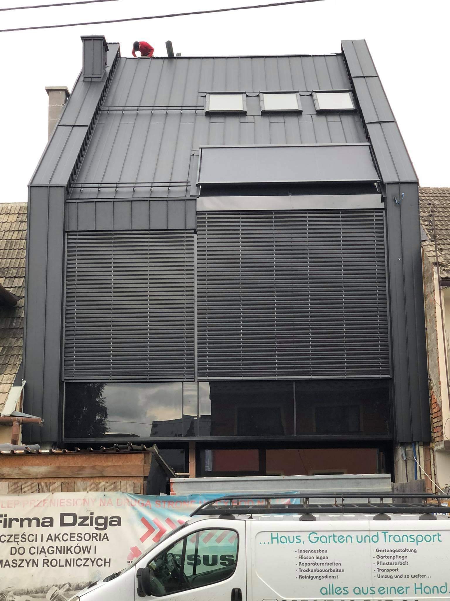 Nowoczesny dom z blachy trapezowej w kolorze grafitowym, okna z żaluzjami zewnętrznymi, dekarz w czerwonej kurtce na dachu, widoczne obróbki blacharskie i komin.