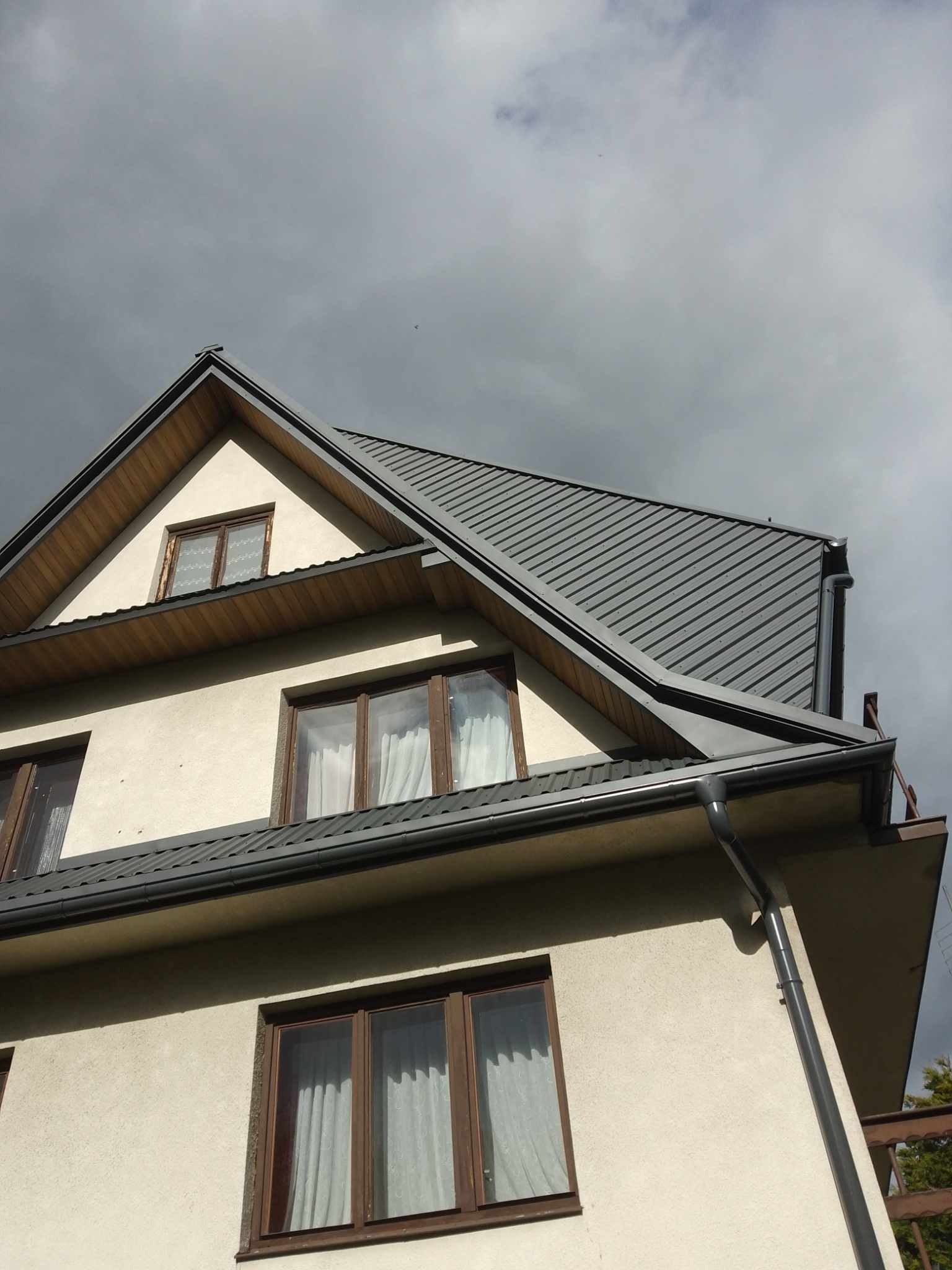Szary blaszany dach z rynnami na dwupiętrowym domu z beżową elewacją i drewnianymi oknami, widok z dołu na tle pochmurnego nieba.