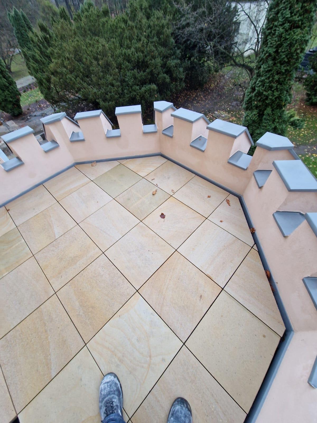 Widok z góry na wykończony taras na dachu w stylu zamkowym, wyłożony kwadratowymi płytami w odcieniach beżu, otoczony murem z blankami, z widocznymi butami osoby robiącej zdjęcie.