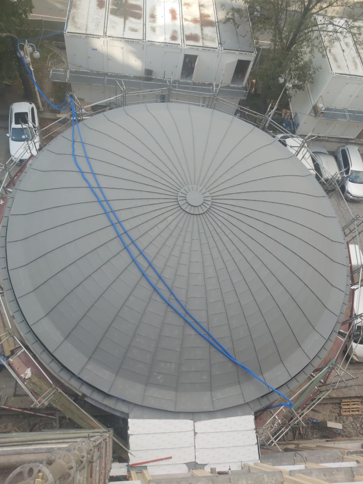 Widok z góry na kopułę pokrytą szarą blachą trapezową, z widocznymi rusztowaniami i niebieskim wężem doprowadzającym wodę, sugerujący prace wykończeniowe.