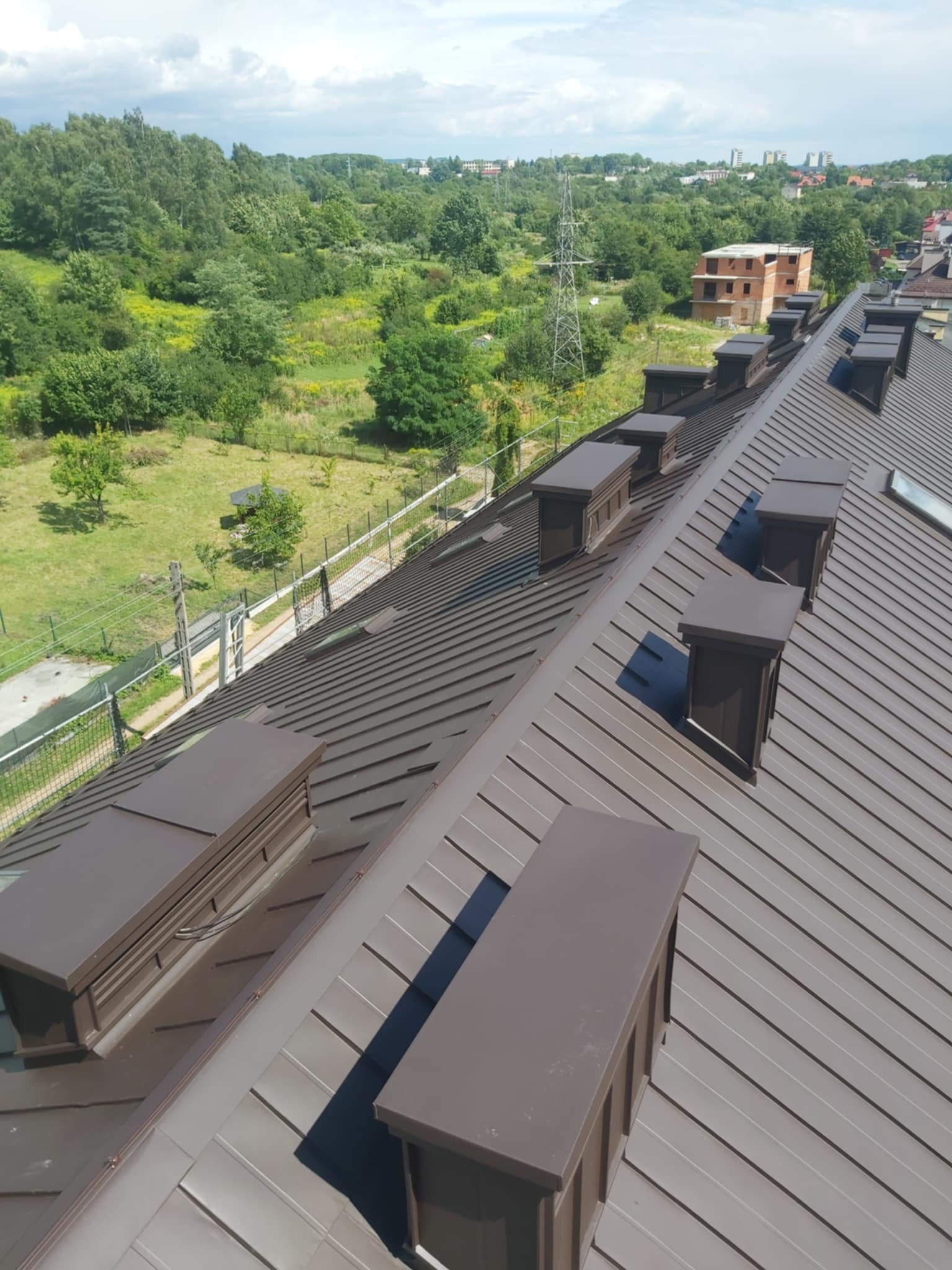 Widok z góry na brązowy, metalowy dach z licznymi kominami wentylacyjnymi, z widocznym krajobrazem zieleni i budynkami w tle.