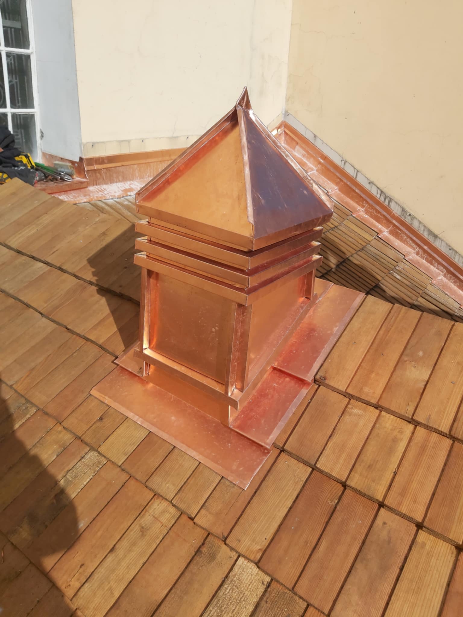 Miedziana, piramidalna obudowa komina na dachu pokrytym drewnianym gontem, widoczny fragment okna i narzędzia dekarskie.
