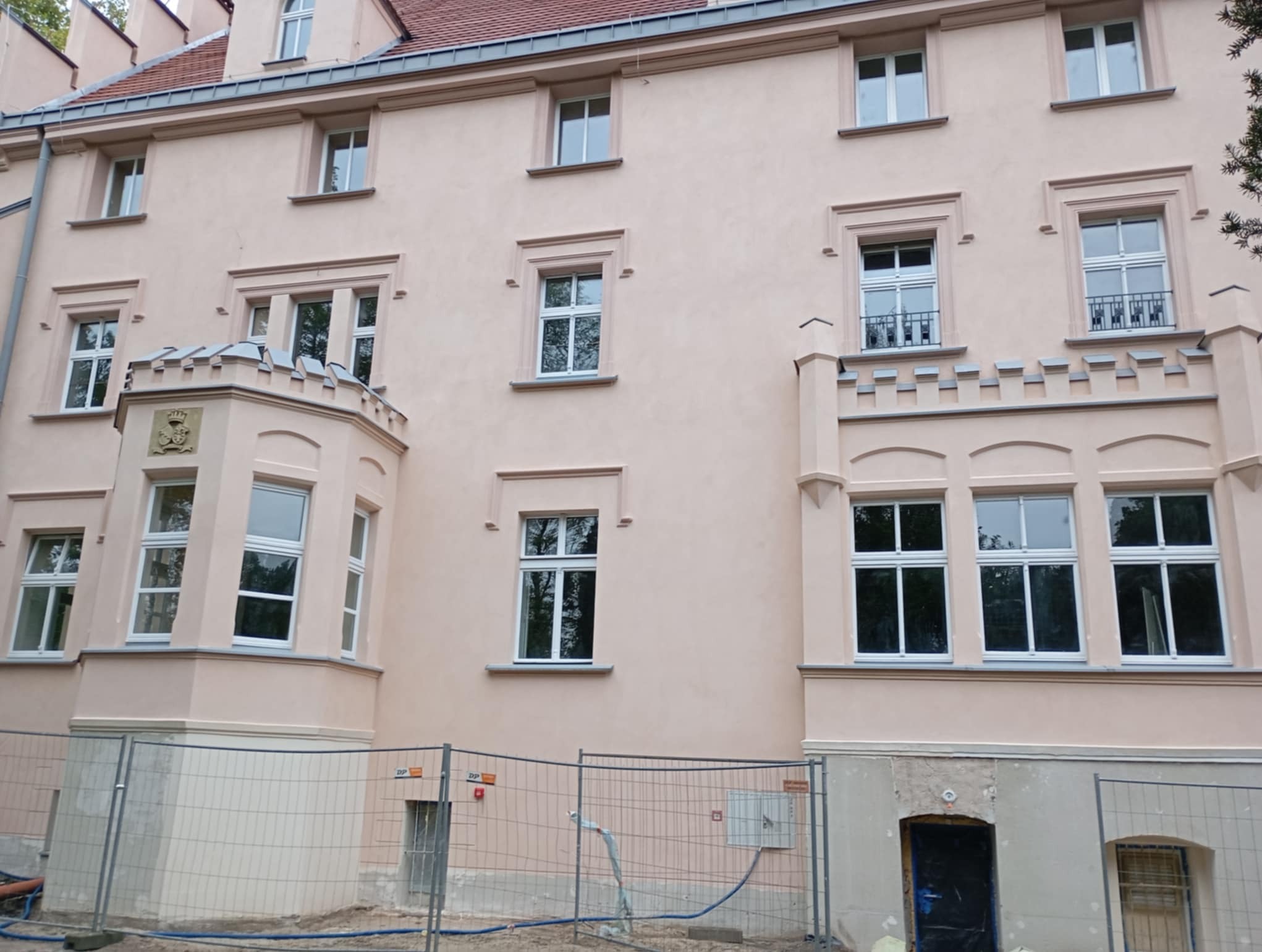 Odnowiona elewacja budynku wielorodzinnego w odcieniach beżu z licznymi oknami o białych ramach, widoczna metalowa siatka zabezpieczająca teren budowy.