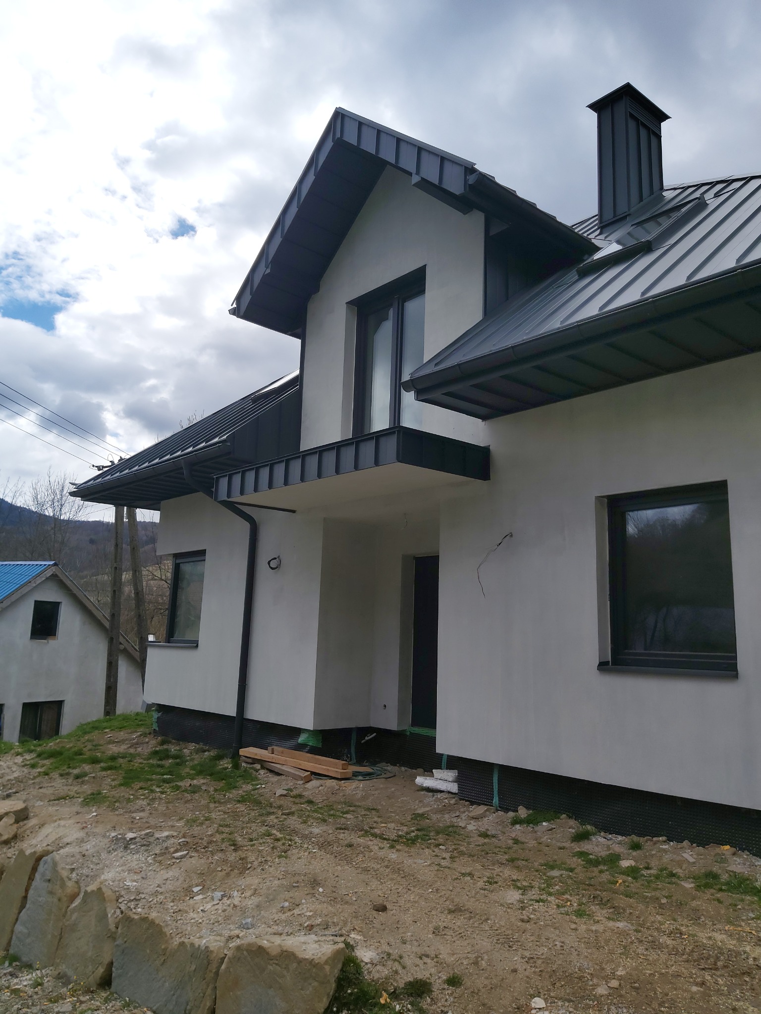 Nowoczesny dom z ciemną, metalową dachówką i rynnami, widoczny komin, elewacja w kolorze złamanej bieli, teren wokół domu w trakcie zagospodarowywania.