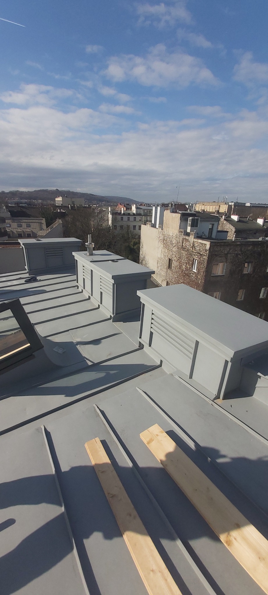 Szary dach z blachy trapezowej z trzema lukarnami wentylacyjnymi i dwoma deskami, widok na zabudowę miejską w tle.