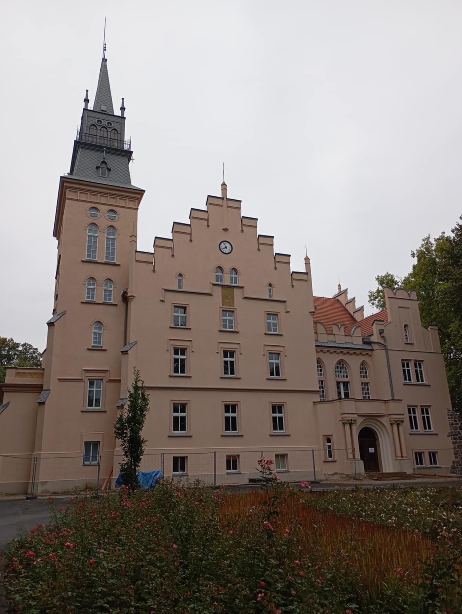 Odnowiony budynek w stylu zamkowym z wieżą, zegarem na fasadzie i czerwonym dachem, otoczony rabatami kwiatowymi i tymczasowym ogrodzeniem.