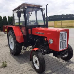 Czerwony traktor Ursus C-360 z kabiną, stojący na kostce brukowej, z widoczną tabliczką znamionową i białymi felgami.