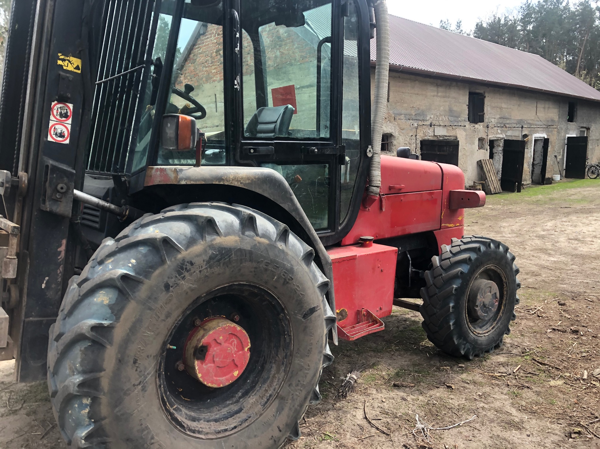 Czerwony wózek widłowy teleskopowy na tle starego, murowanego budynku gospodarczego z ceglanym dachem.