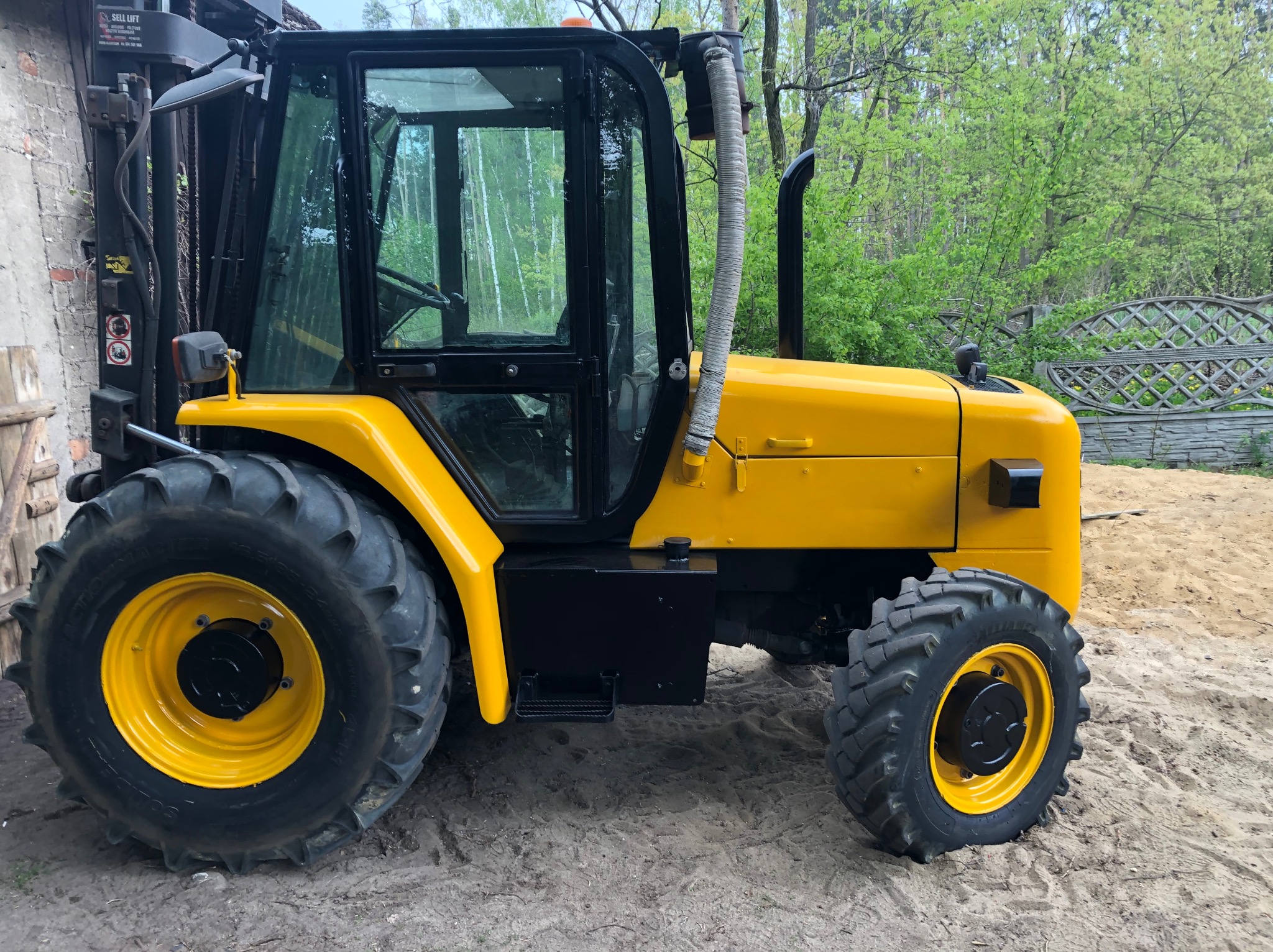 Żółty ciągnik z czarną kabiną i dużymi oponami stoi na piaszczystym podłożu, widoczny fragment budynku i zielone drzewa w tle.