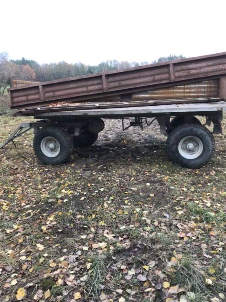 Przyczepa rolnicza z metalową skrzynią, widoczne ślady korozji, stojąca na trawie pokrytej opadłymi liśćmi, tło stanowią drzewa.
