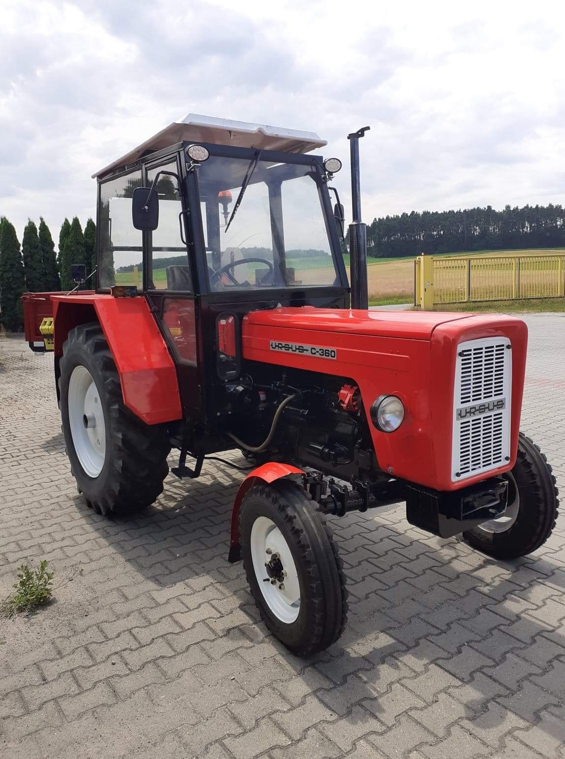Czerwony traktor Ursus C-360 z kabiną, stojący na kostce brukowej, z widoczną tabliczką znamionową i białymi felgami.