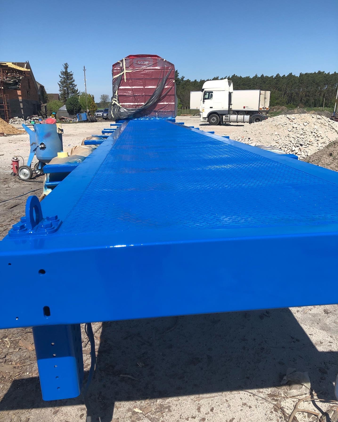 Świeżo pomalowana na niebiesko naczepa samochodowa z widoczną fakturą antypoślizgową, obok maszyna do piaskowania, w tle ciężarówka i gruzowisko.