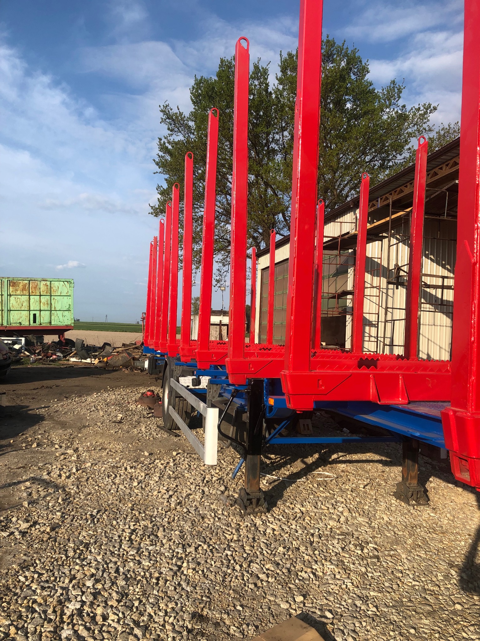 Czerwona, nowo pomalowana przyczepa do transportu drewna z podniesionymi kłonicami, stojąca na żwirowym placu, w tle zielony kontener i budynek gospodarczy