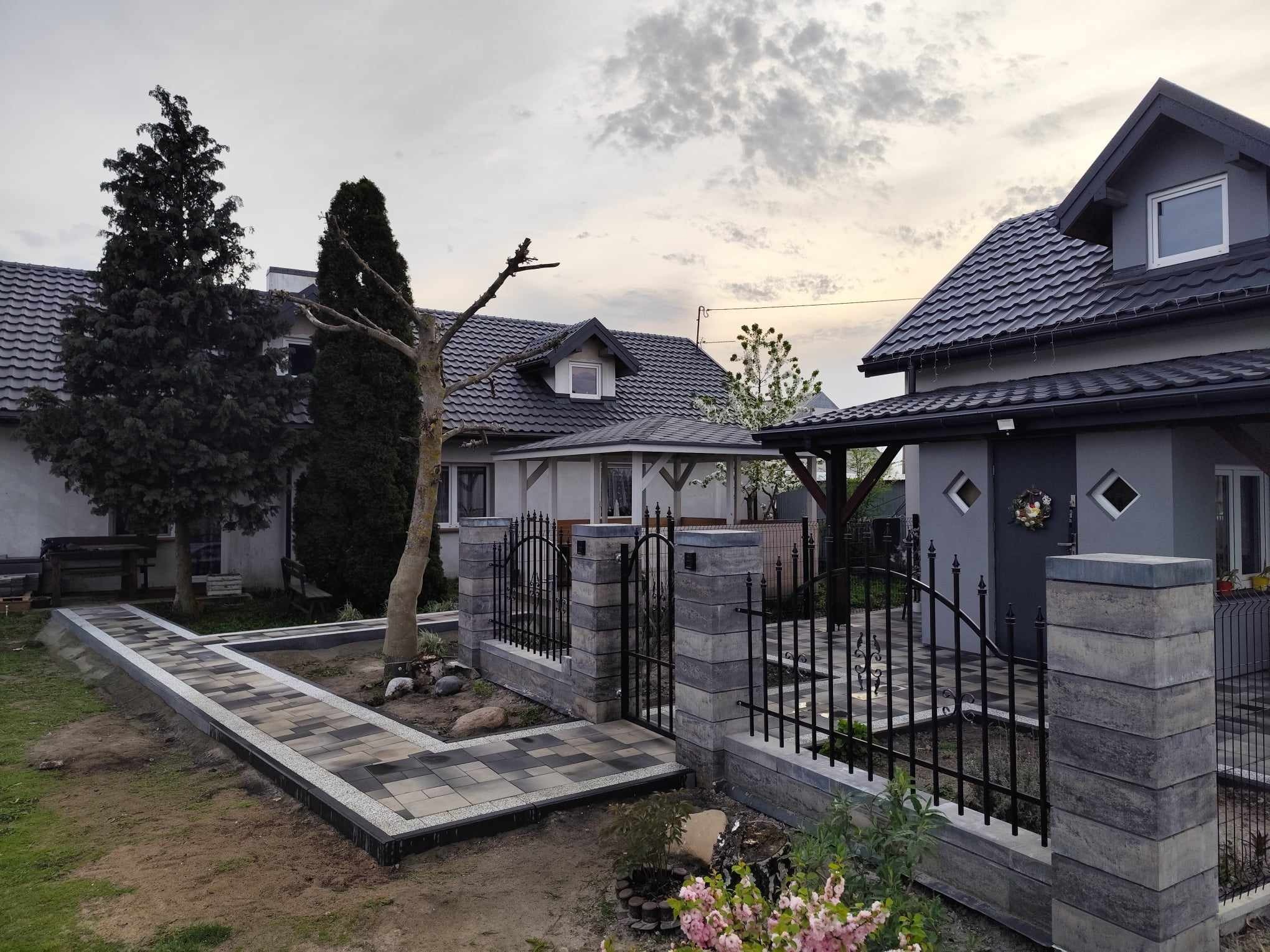 Nowoczesna elewacja domu z szarą kolorystyką, czarną dachówką i elementami dekoracyjnymi, otoczona zadbanym ogrodem z ścieżką z kostki brukowej i czarnym ogrodzeniem.