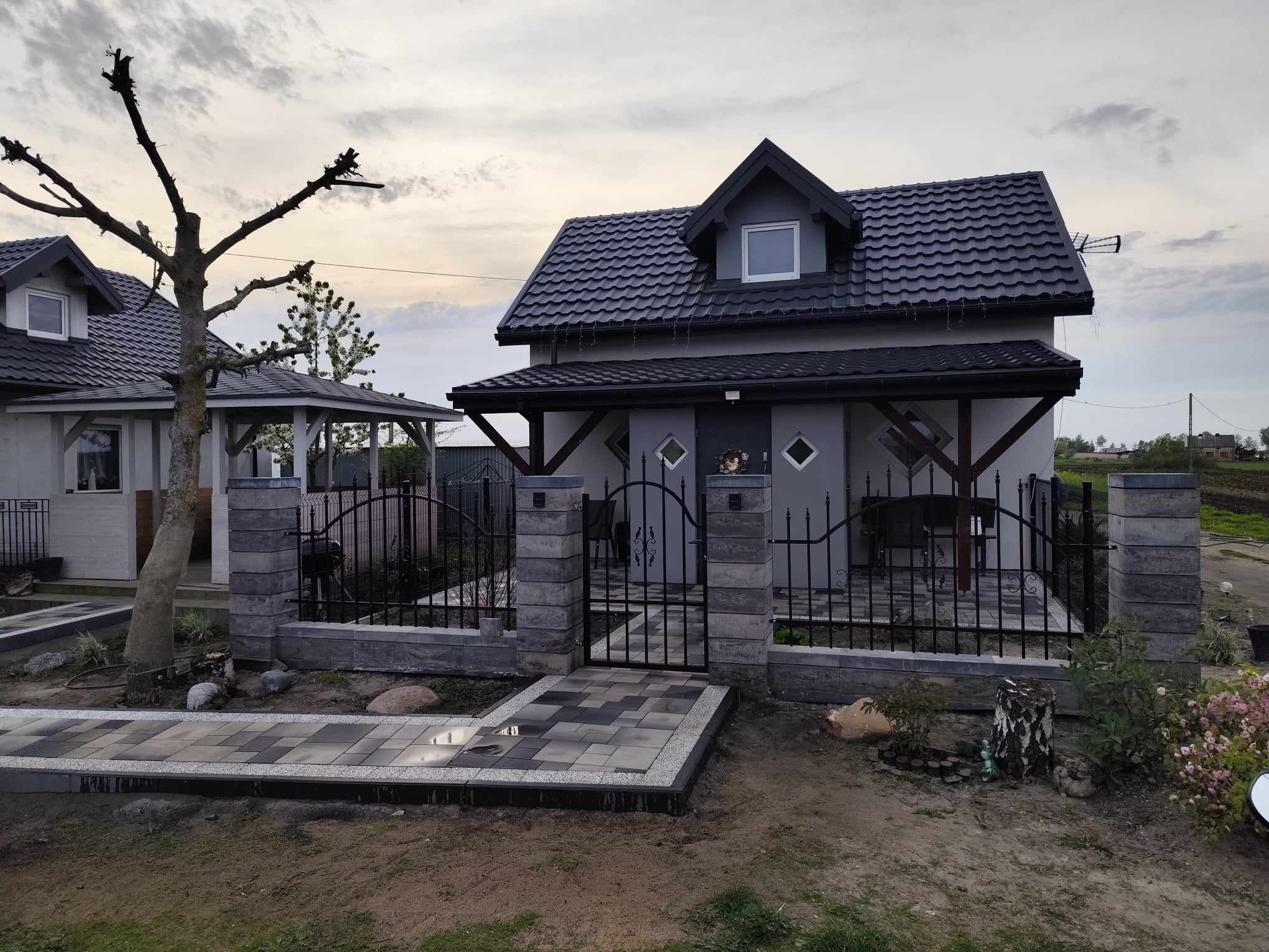 Szary dom z czarnym dachem i ogrodzeniem z kamiennych słupków. Ułożona kostka brukowa prowadzi do furtki. Widoczny fragment drugiego budynku i drzewo bez liści.