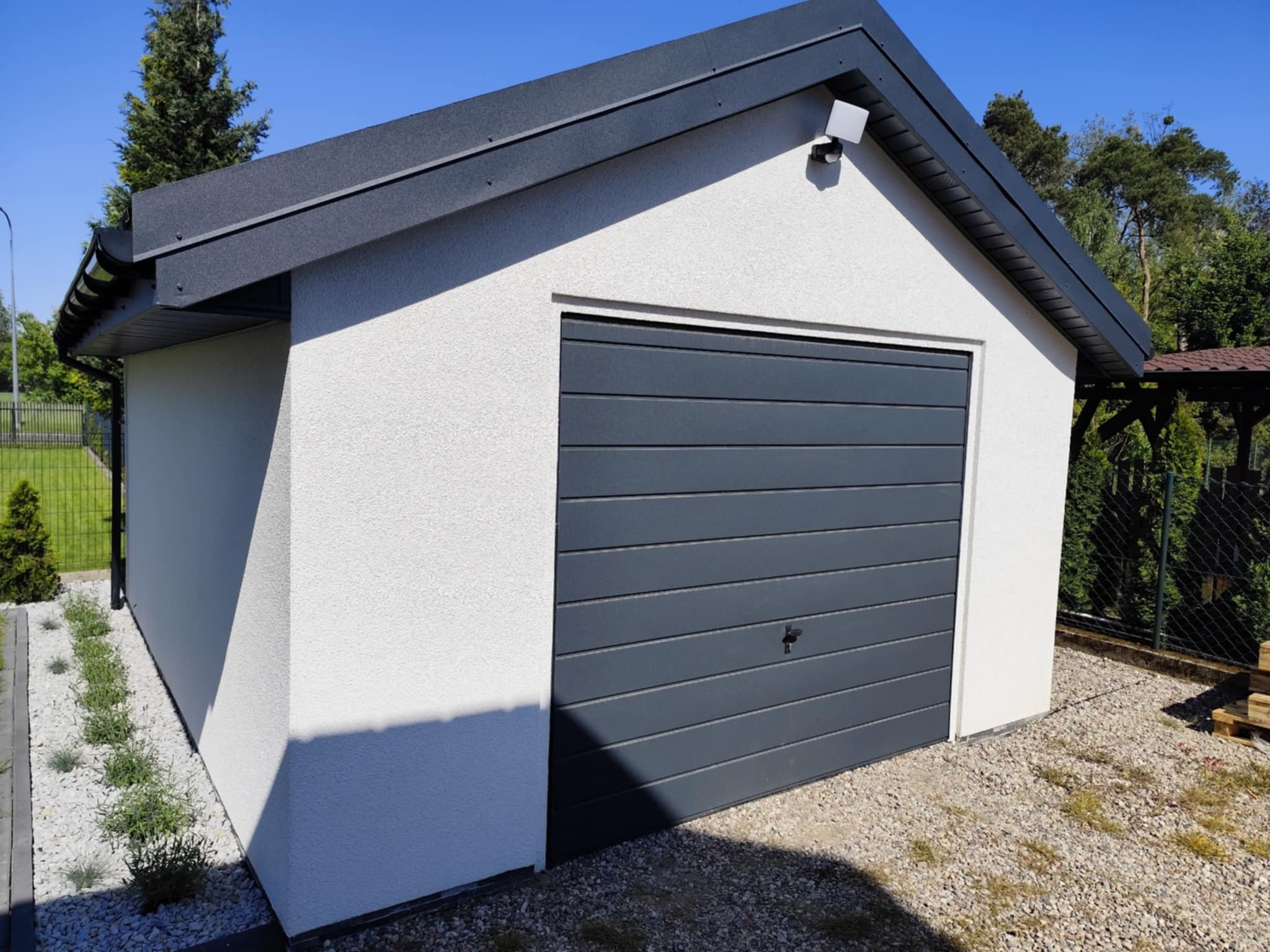 Nowoczesny garaż z elewacją w kolorze złamanej bieli i szarą bramą segmentową, otoczony zielenią w Obrowie. Widoczne oświetlenie zewnętrzne i wykończenie dachu.