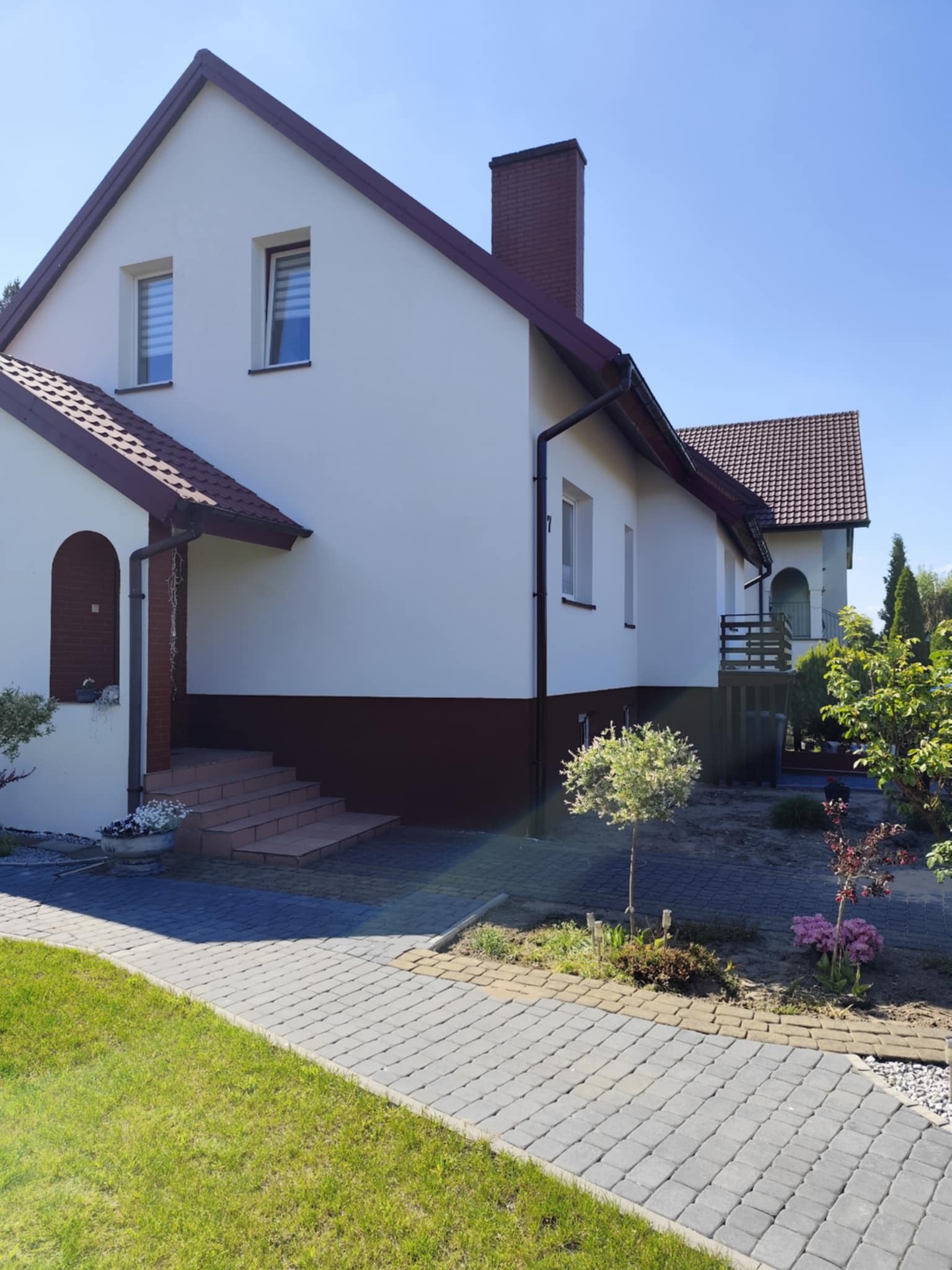 Dom jednorodzinny z jasną elewacją i ciemnobrązowym akcentem u dołu, widok z chodnika z kostki brukowej, ogród z niskimi drzewkami i kwiatami.