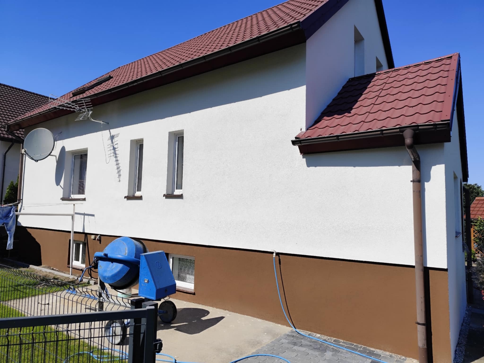 Dwukolorowa elewacja domu z brązowym cokołem i białą górą. Na pierwszym planie betoniarka, sugerująca prace wykończeniowe. Widok z niskiej perspektywy.