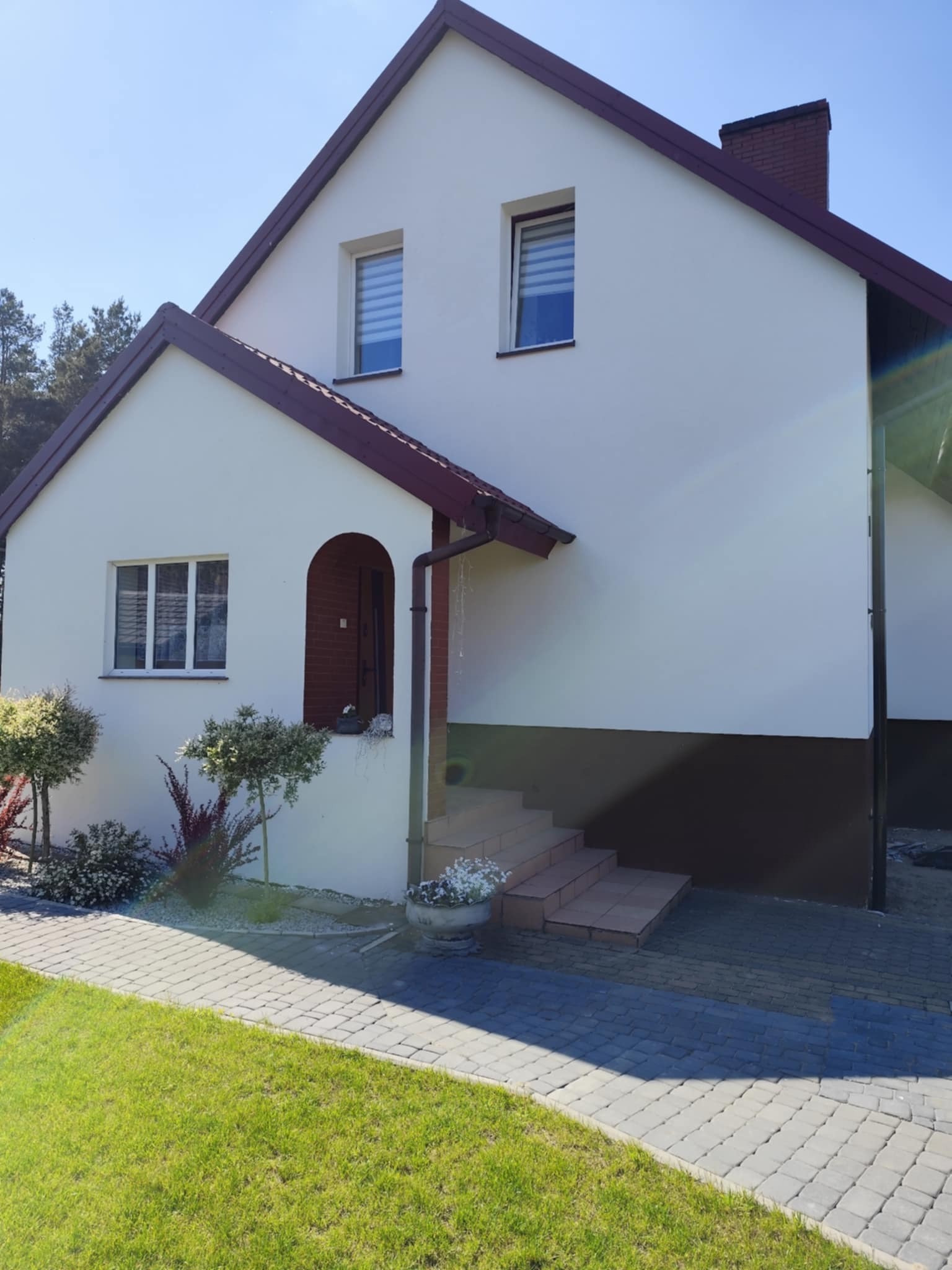 Dwukondygnacyjny dom z jasną elewacją i brązowym dachem, widok z poziomu trawnika. Wejście z łukowym zadaszeniem, schody z terakoty. W tle drzewa.