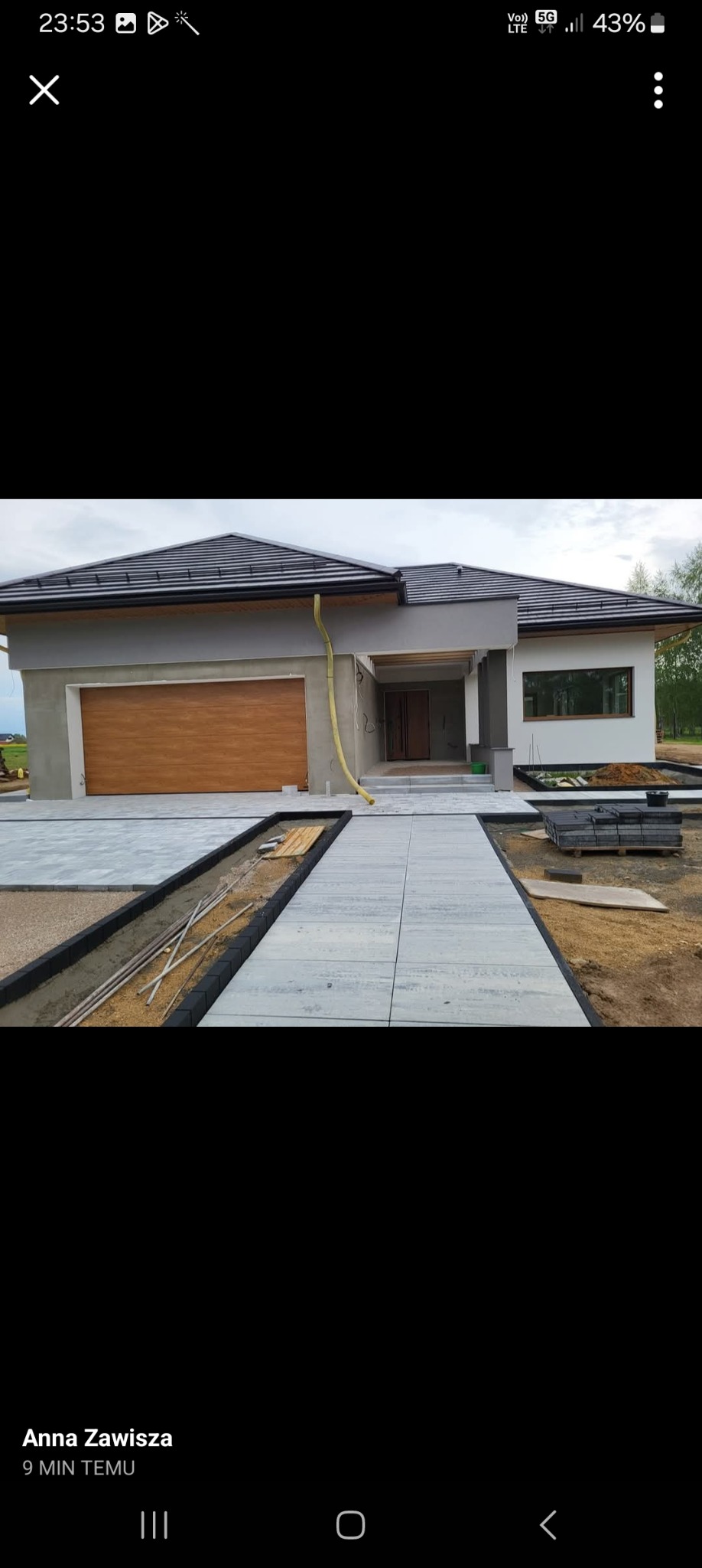 Nowoczesny dom w trakcie budowy: szara elewacja, brązowa brama garażowa, ułożona kostka brukowa na podjeździe. Częściowo wykończona elewacja i zagospodarowanie terenu.